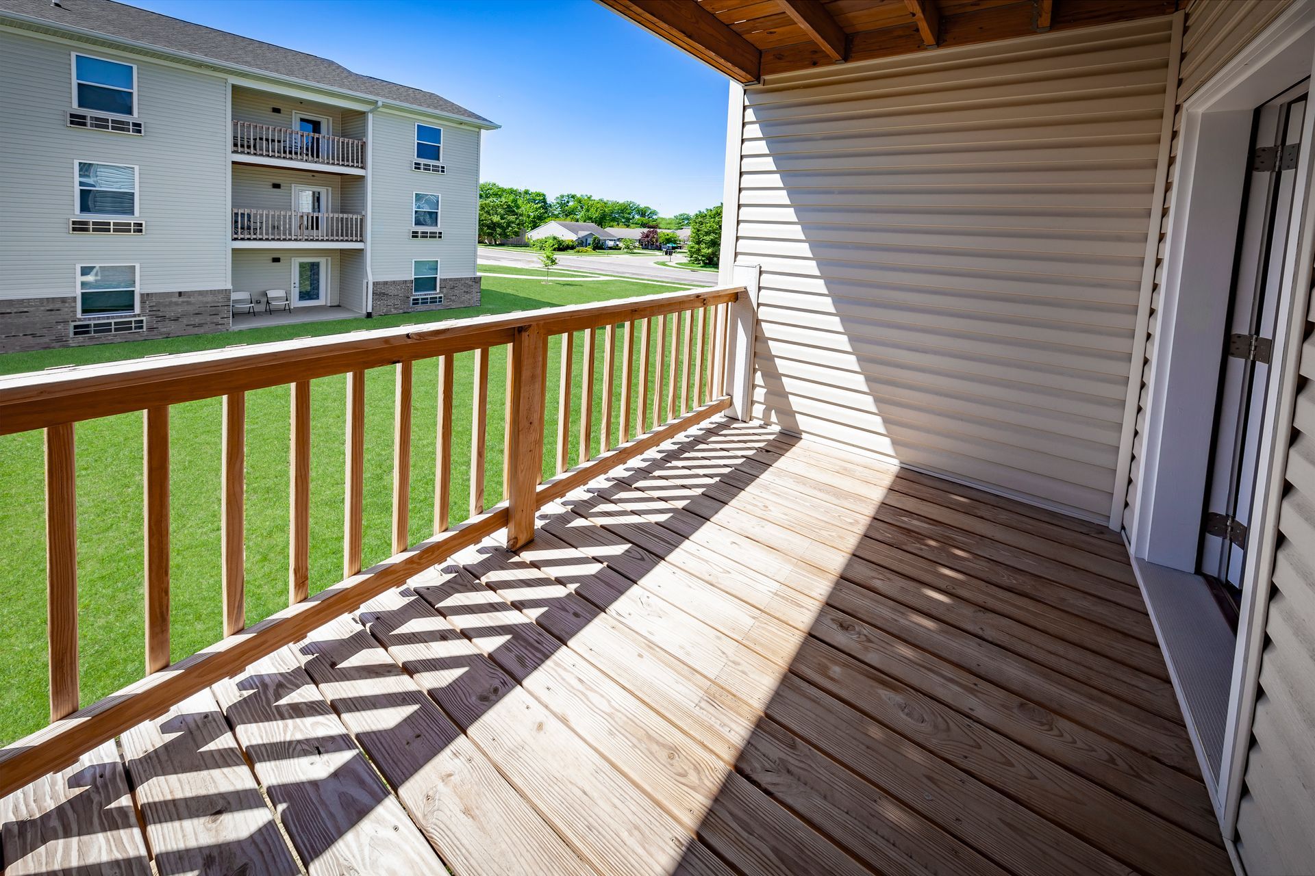 Apartment balcony with wooden railing and sunlit deck overlooking a grassy courtyard