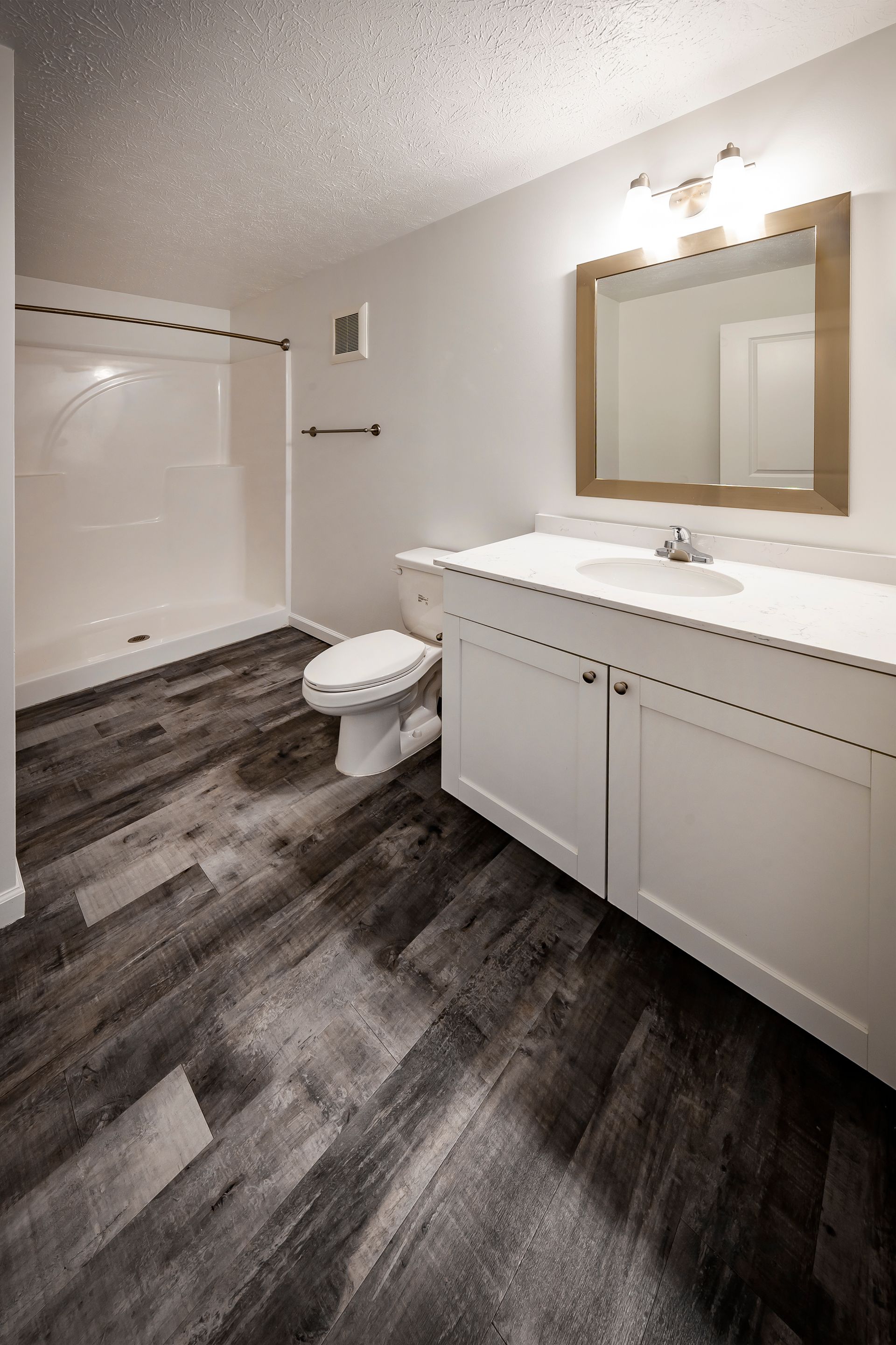 Modern bathroom with white vanity, large mirror, tub-shower, toilet, and dark wood-look flooring.