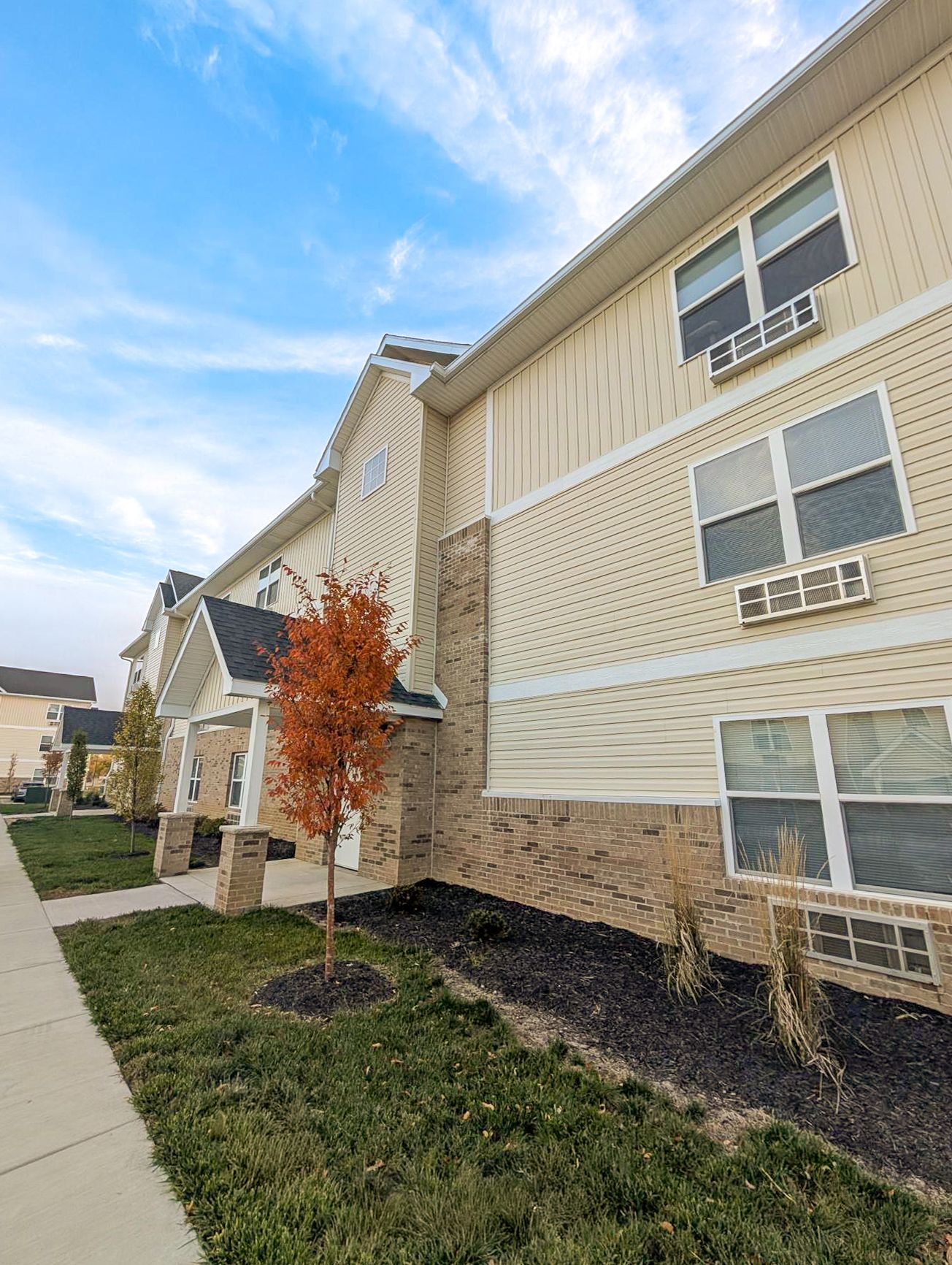 Two-story beige apartment building with balconies, set against a cloudy sky.
