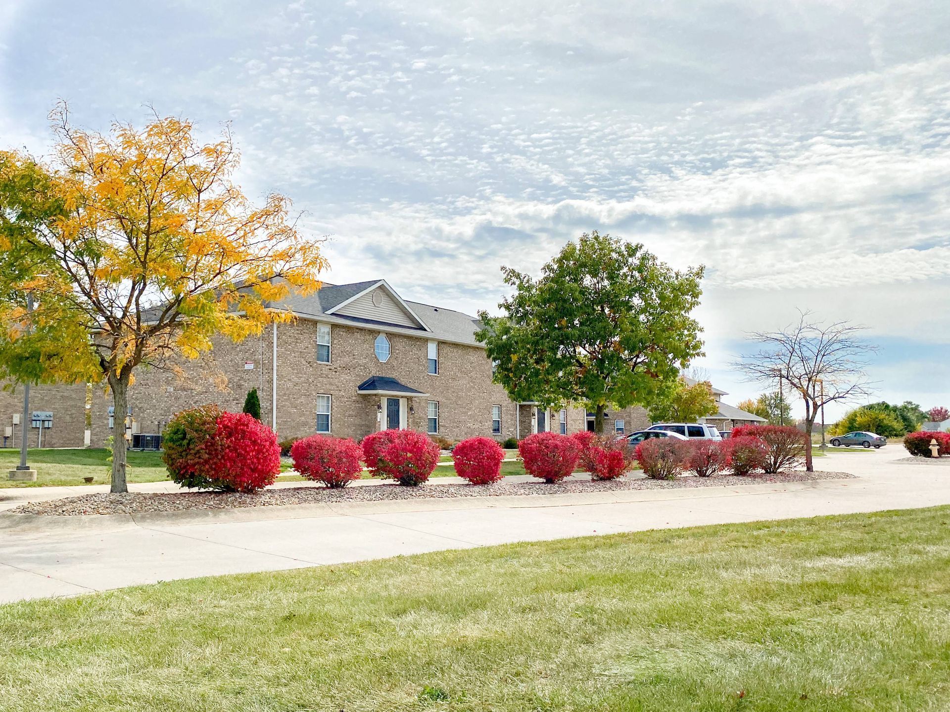 Brick building with colorful autumn trees, red bushes, and a grassy lawn under a cloudy sky.