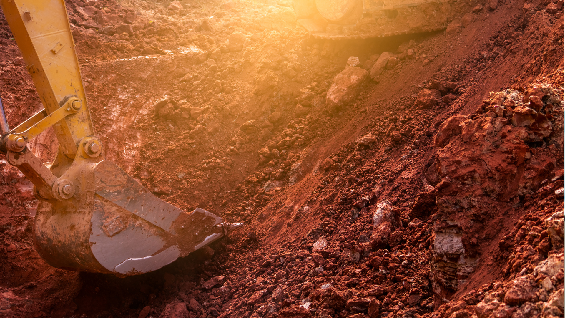 A yellow excavator bucket scoops red soil from a construction site, illuminated by bright sunlight.