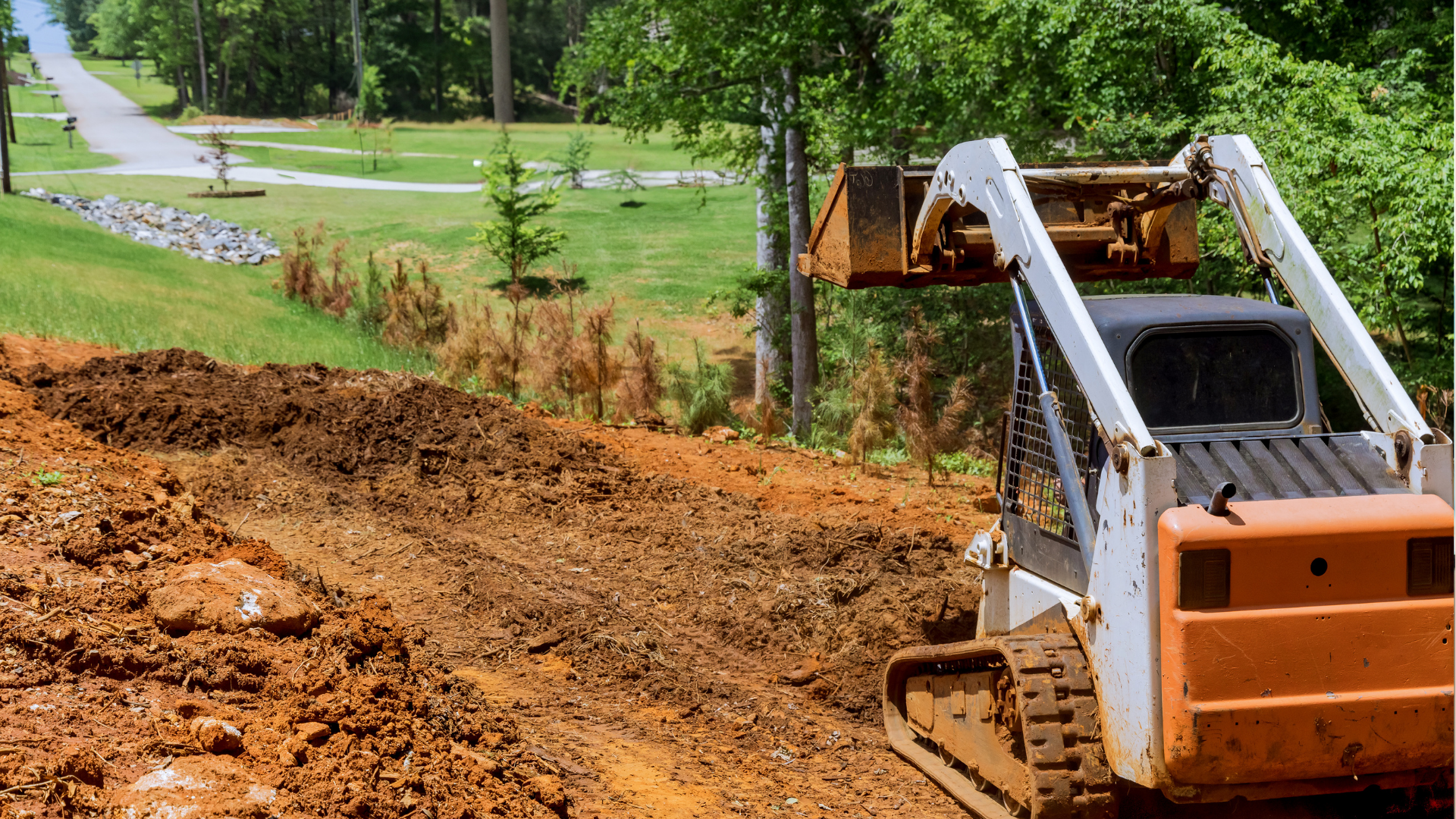 Orange and white skid-steer loader on a dirt hill, moving earth near a road and grassy area.