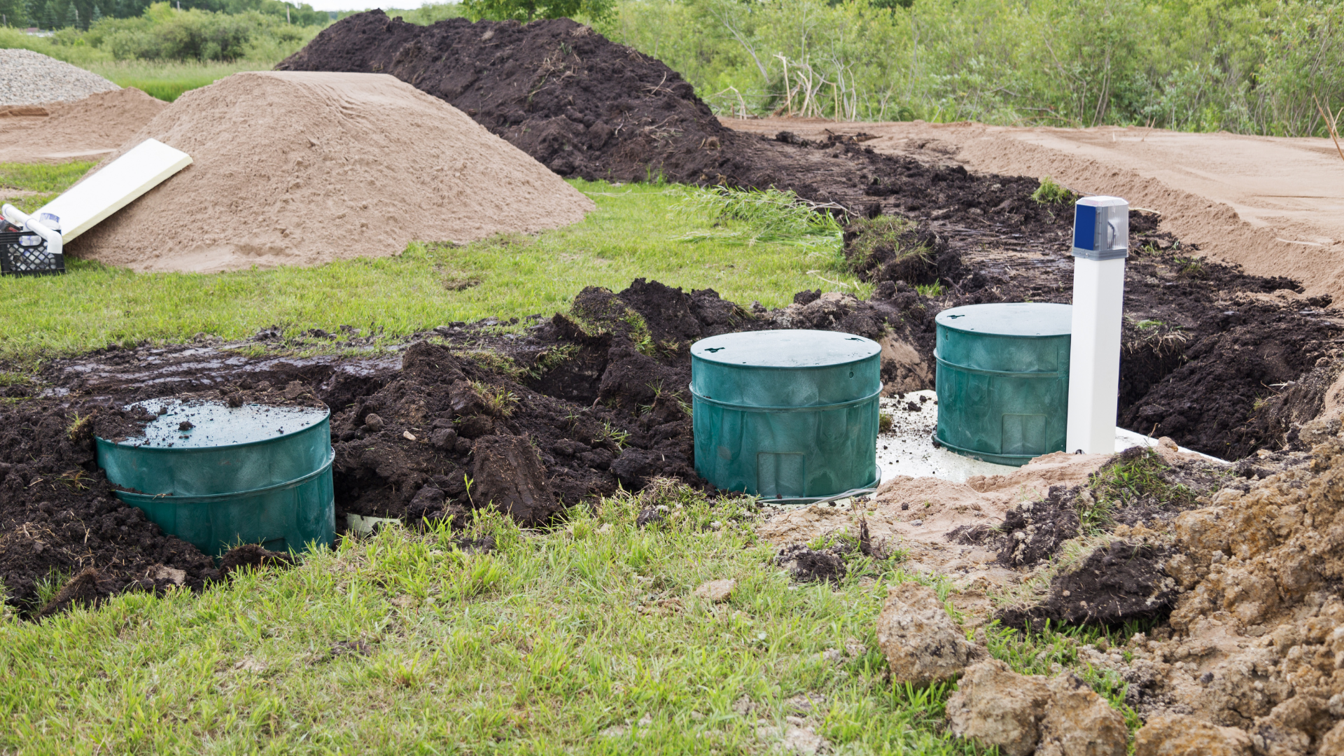 Septic system components installed in a grassy yard, with dirt mounds and a sand pile in the background.