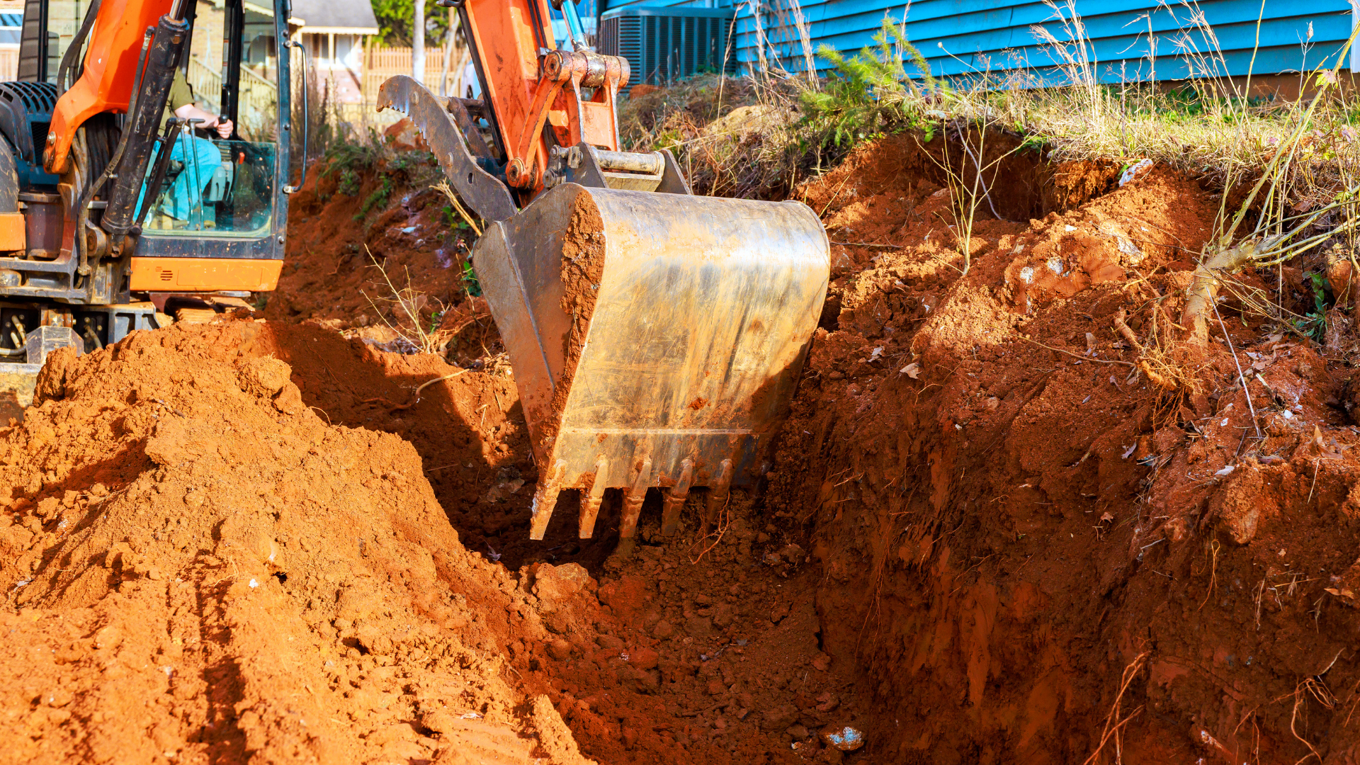 An orange excavator bucket digging a trench in reddish-brown soil on a construction site.