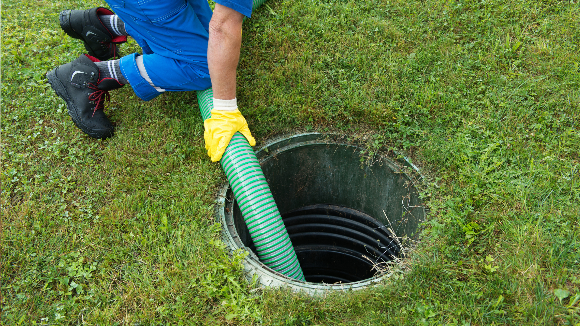 Worker in blue clothes and yellow gloves using a green hose to inspect a ground drain hole in grass.