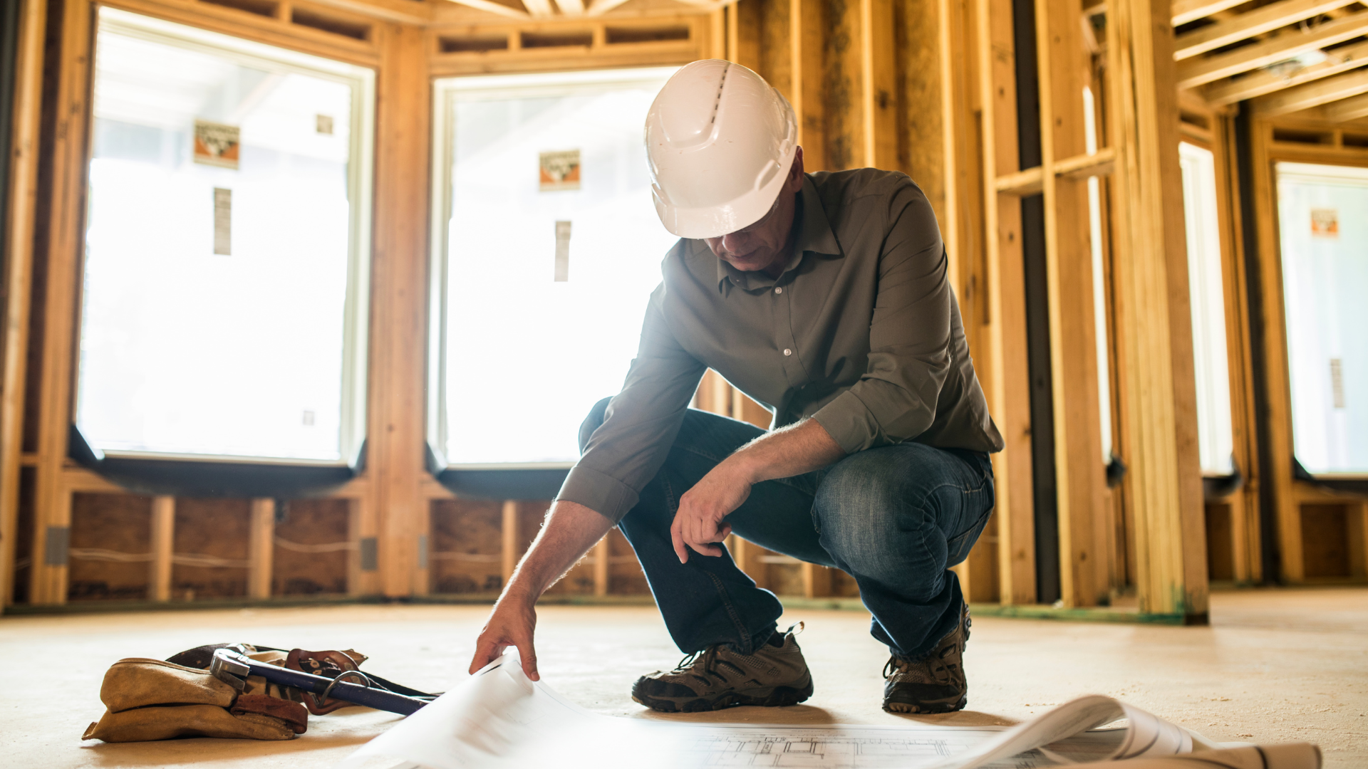 A man is kneeling down in a room in a house under construction looking at a blueprint.