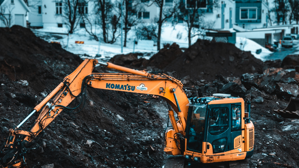 Yellow excavator digging into a pile of dirt and rocks, with a cityscape in the blurred background.