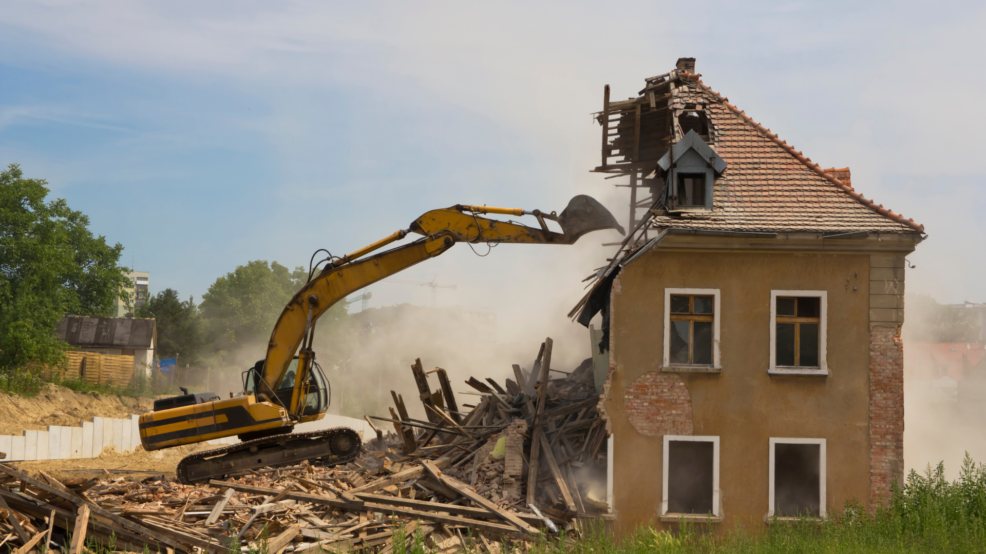 A yellow excavator demolishing the side of a two-story, tan-colored house amidst a cloud of dust.
