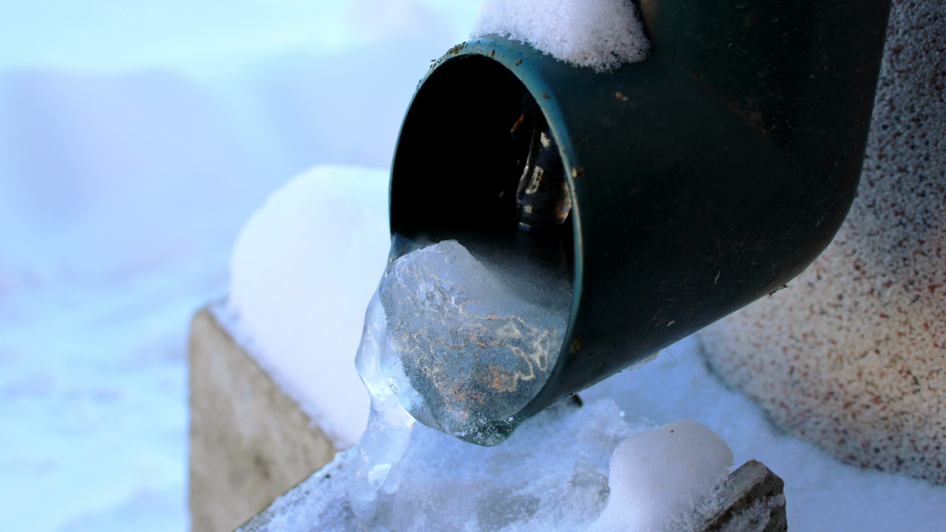 A frozen, green metal downspout dripping with icicles in a snowy outdoor setting.