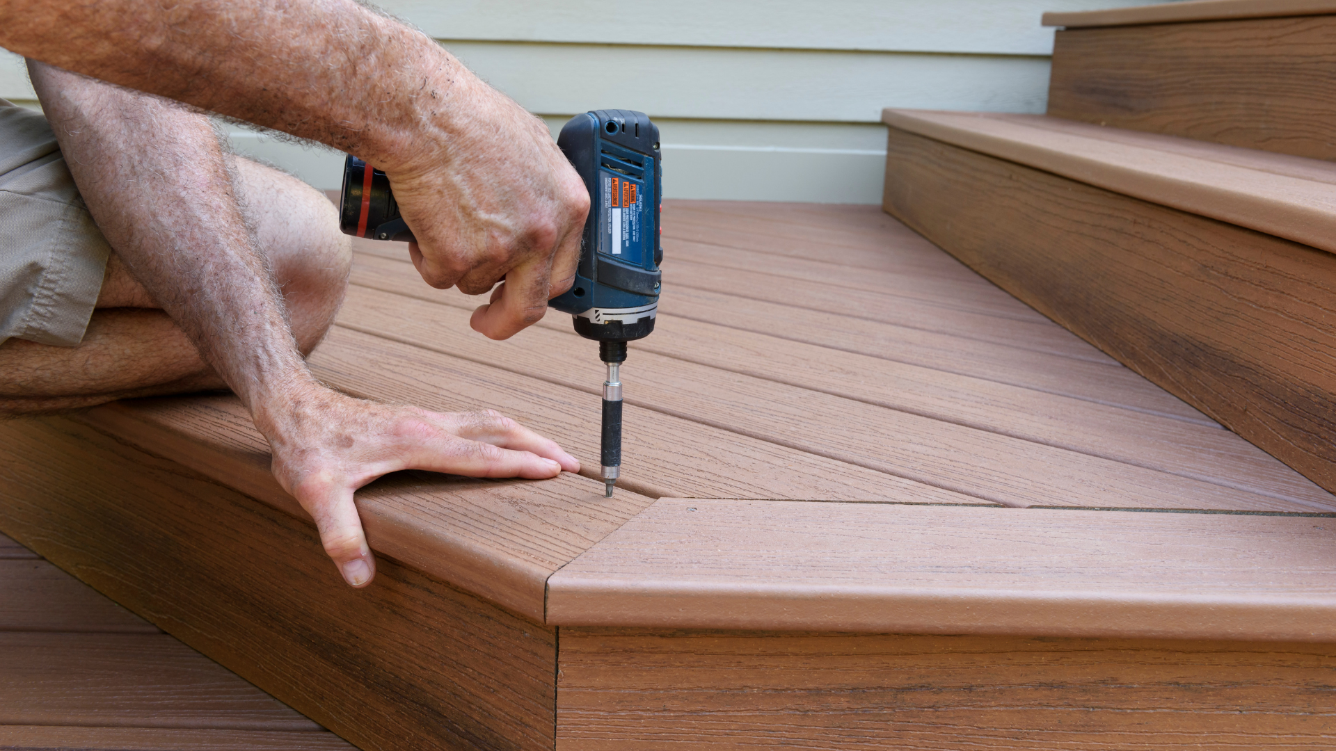 Person using a drill to install a wooden deck on stairs outdoors.