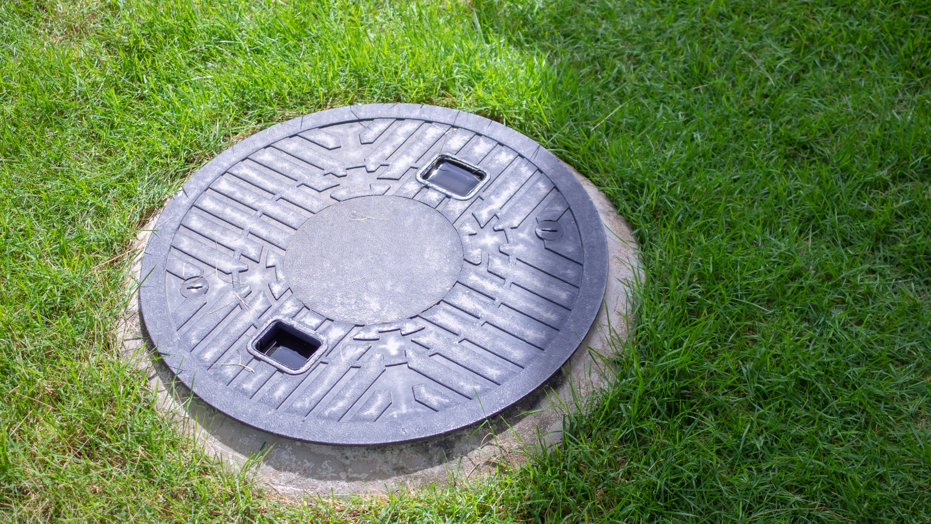 Round, dark septic tank cover in green grass.