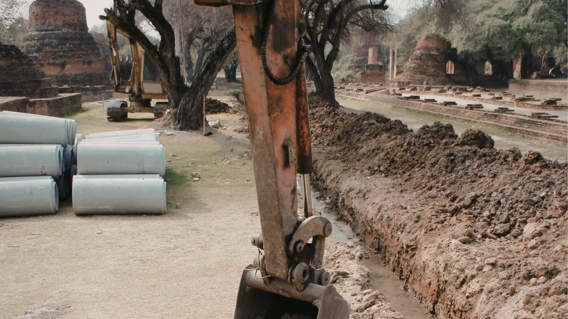 An excavator digging a trench near ancient ruins, with large pipes nearby.