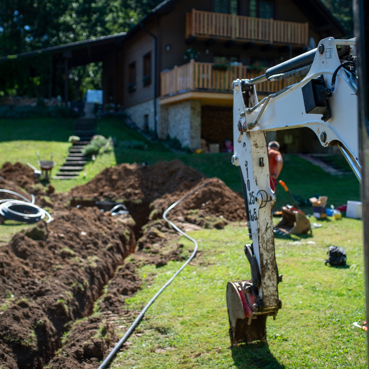 A close-up view of an excavator arm in the foreground, with a dirt trench and a house in the background.