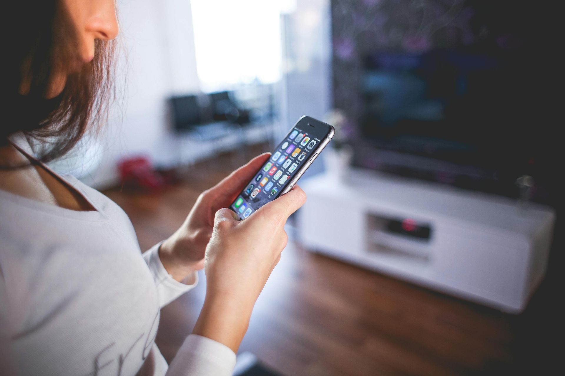 Person holding a smartphone indoors, scrolling. The room has a TV and a white cabinet.