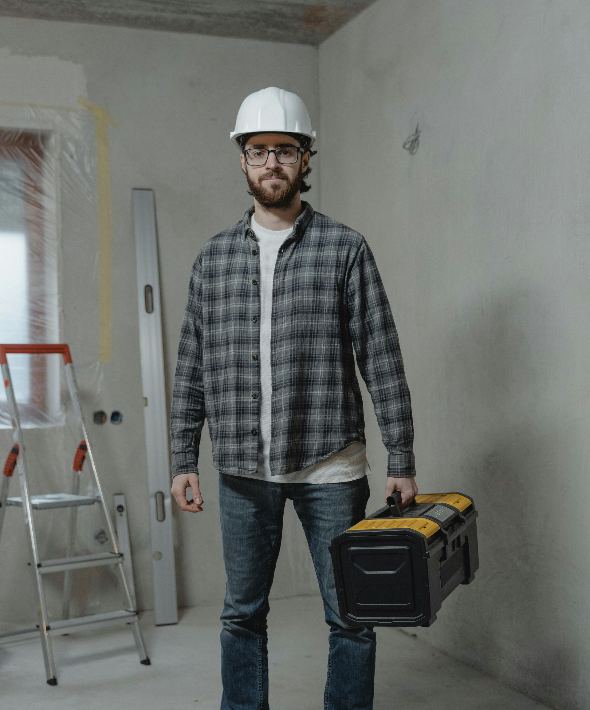Construction worker in hard hat, holding a toolbox, standing in a room under construction, with a ladder.