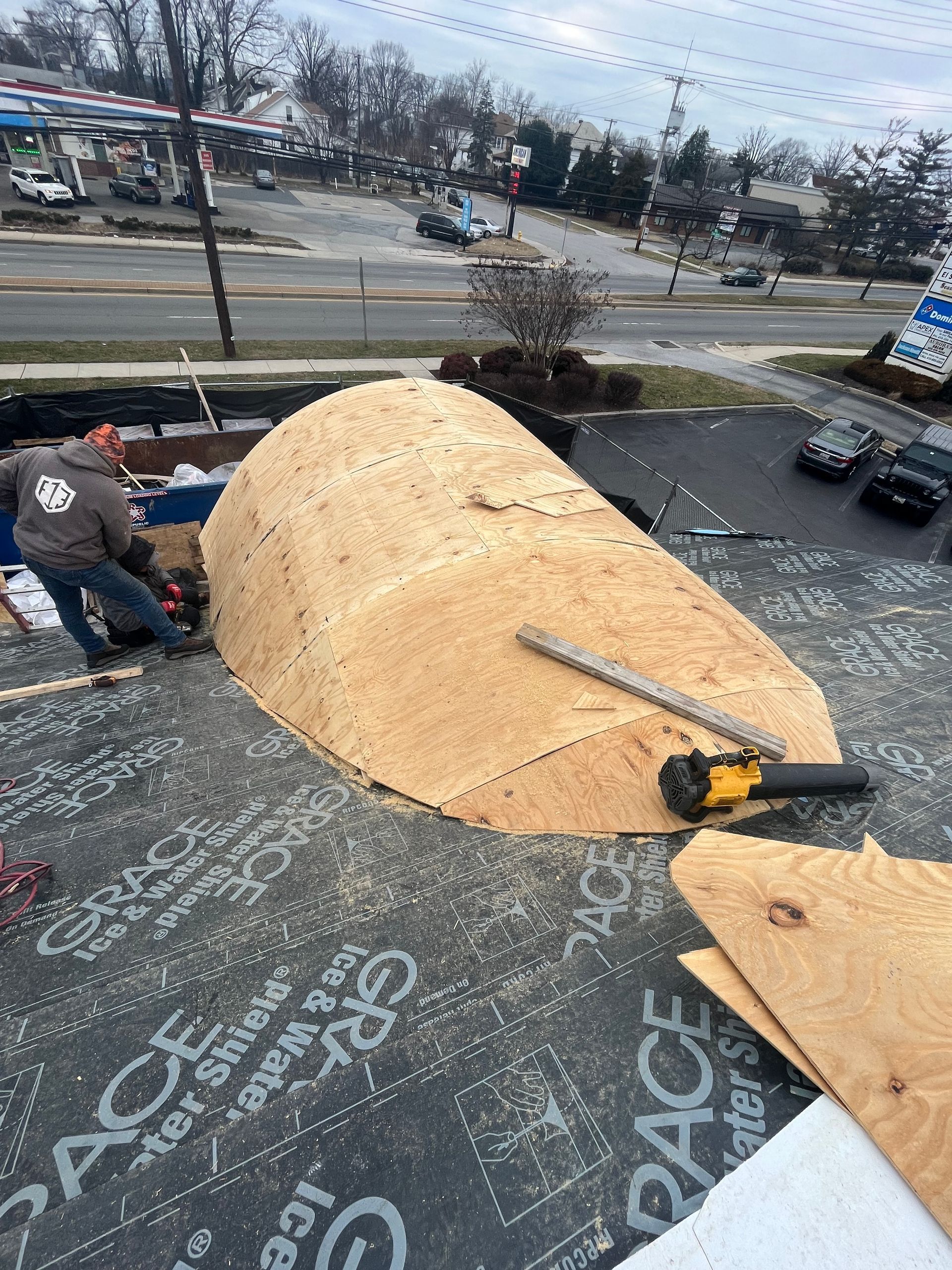 Construction worker on a roof building a wooden dome-like structure, with tools and a busy street in the background.