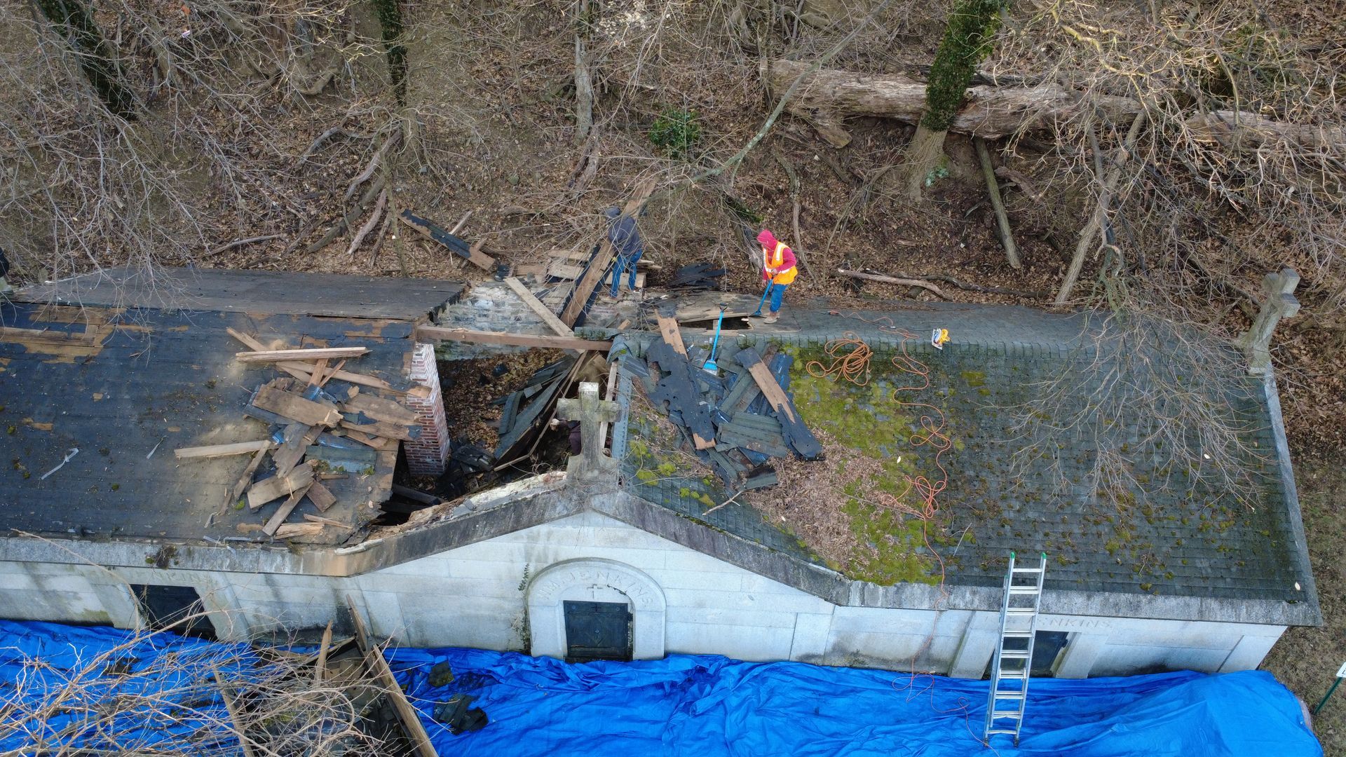 Overhead view of a damaged building with a person in an orange vest on the roof. Blue tarp covers part. Trees in background.