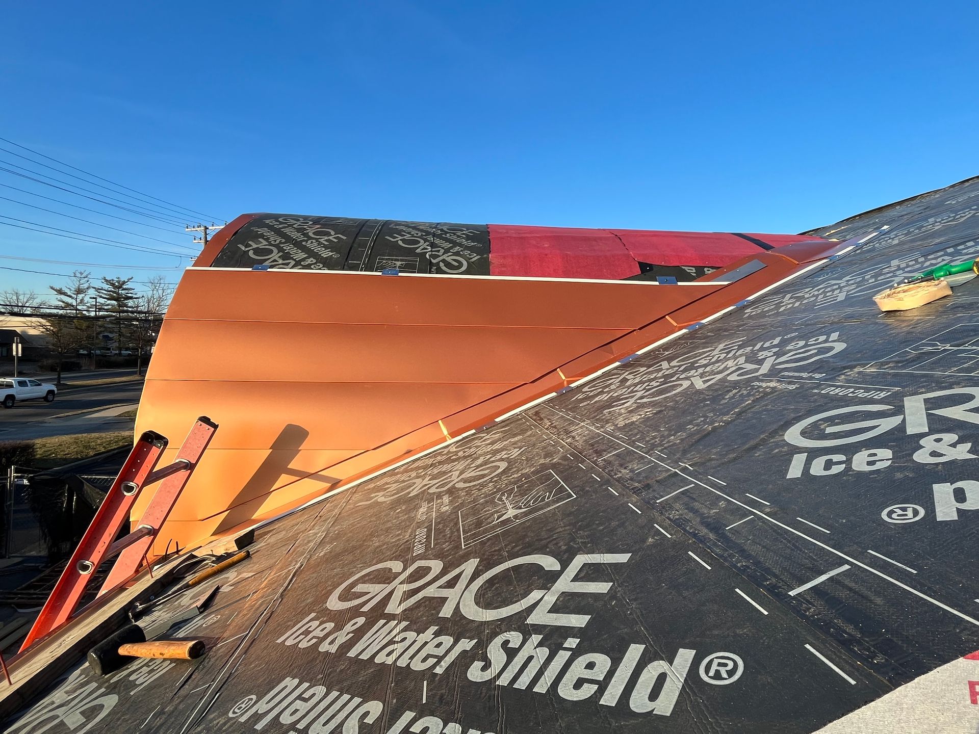 Roof construction: Copper flashing over red underlayment, black water shield, orange ladder, clear sky.