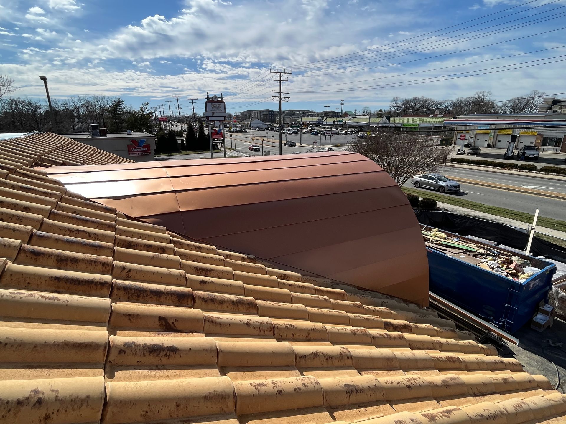 View from a tile roof overlooking a street with buildings, a dumpster, and a blue sky with clouds.