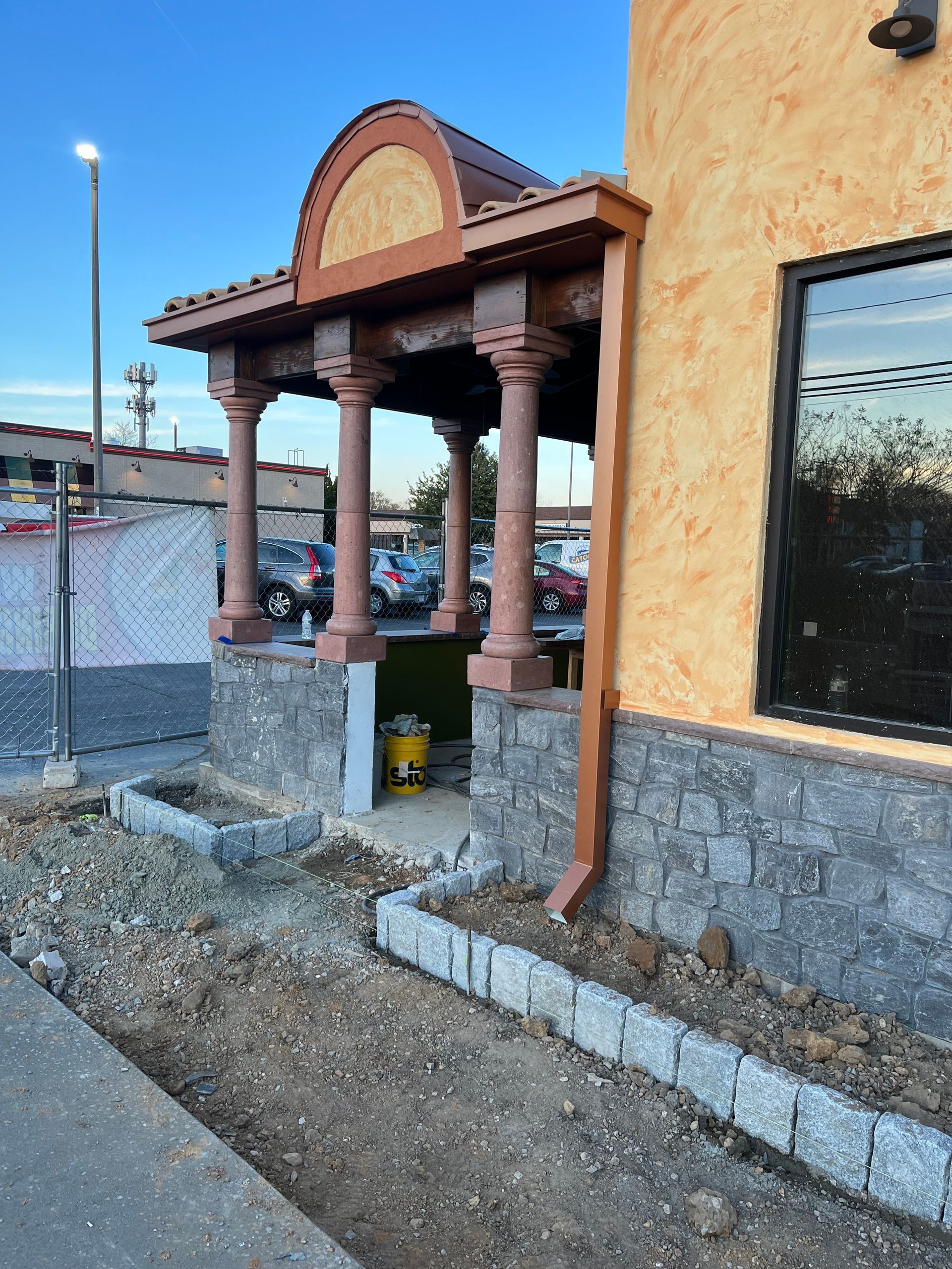 Restaurant entrance with columns, brown roof, and textured orange wall. Gray brick planters and walkway under construction.