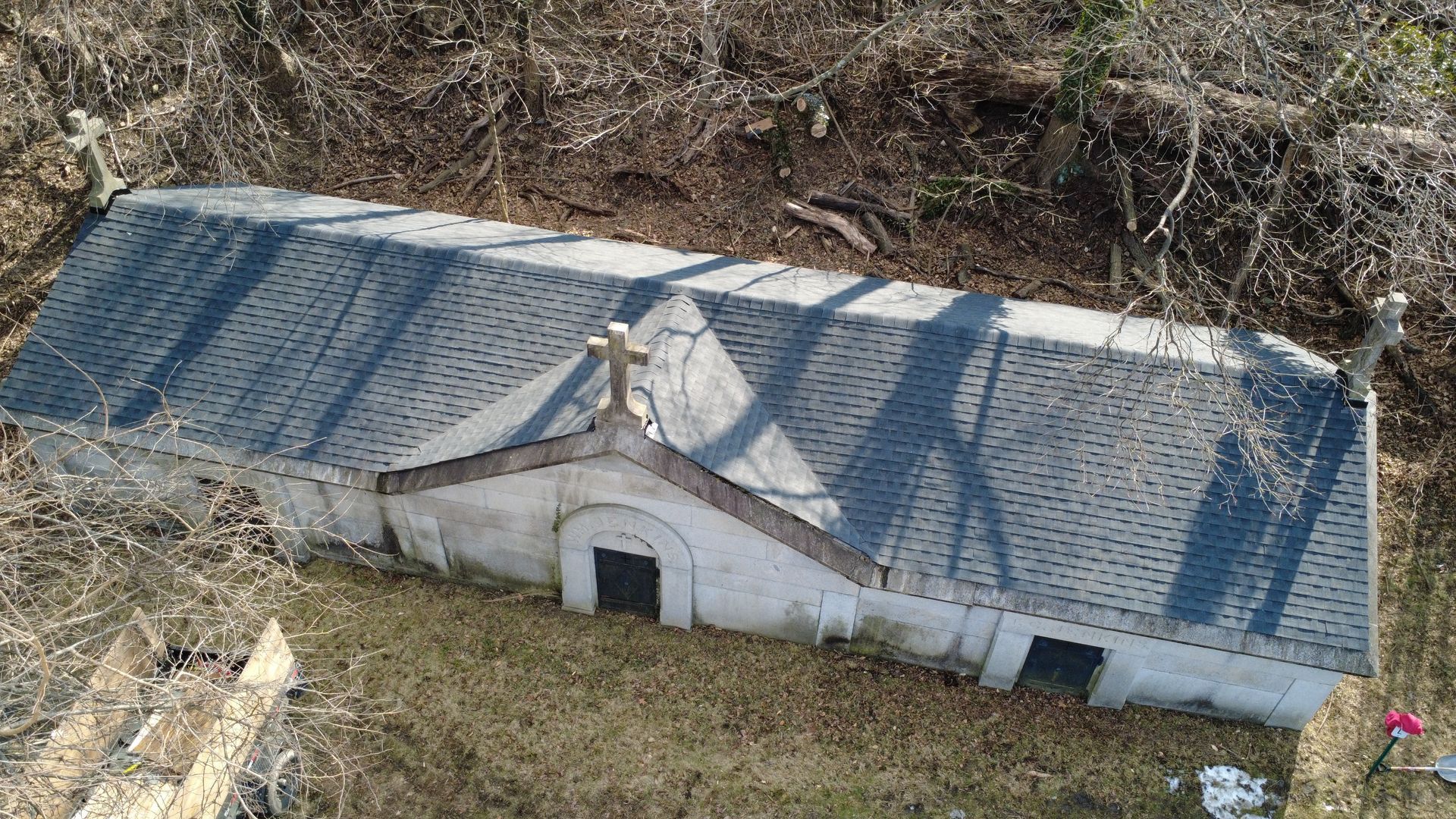 Overhead view of a white, weathered mausoleum with a dark gray shingled roof and crosses. Located in a grassy area.