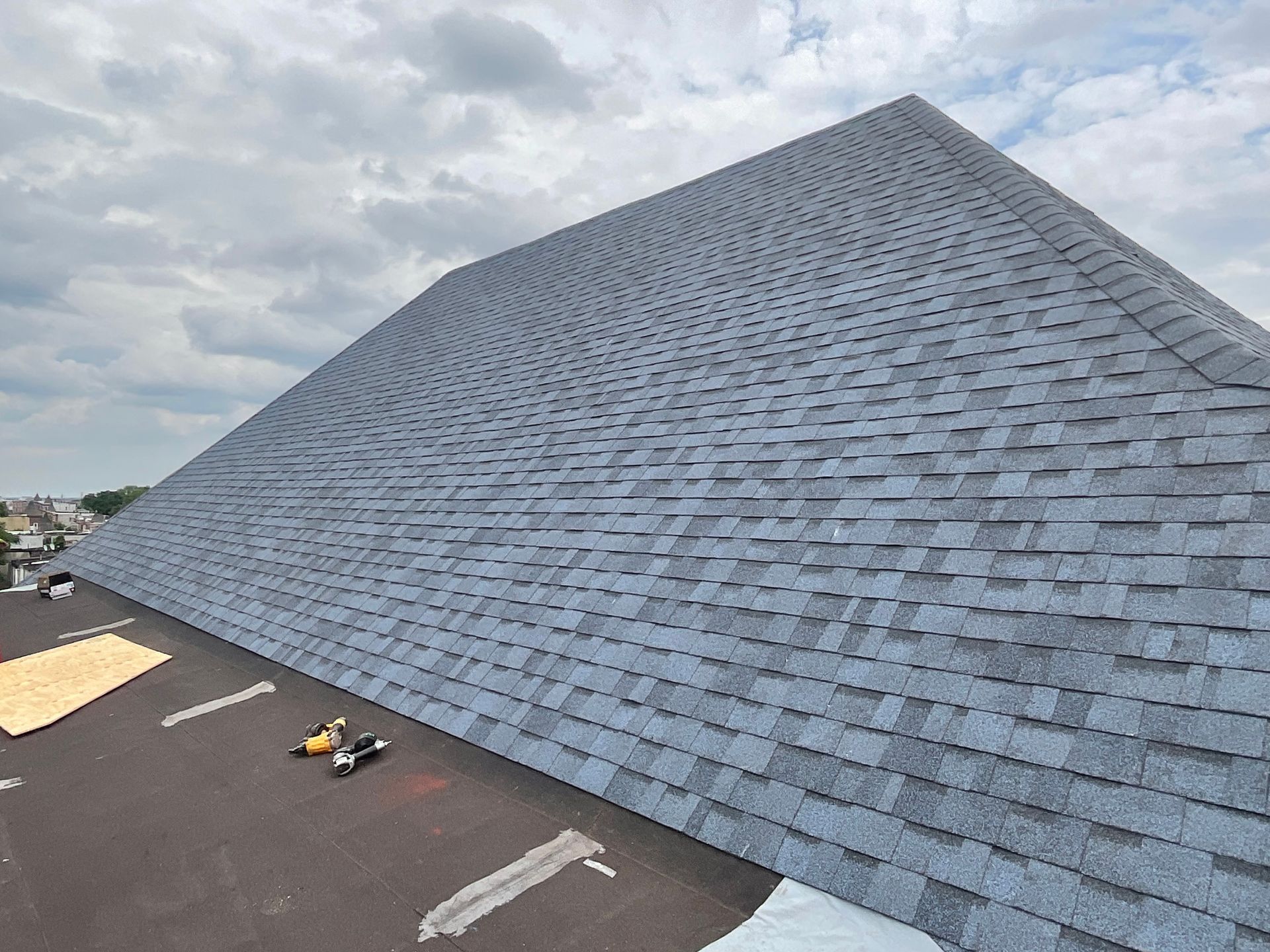 Newly shingled, gray roof on a building under a cloudy sky. Tools and boards are visible on the roof.
