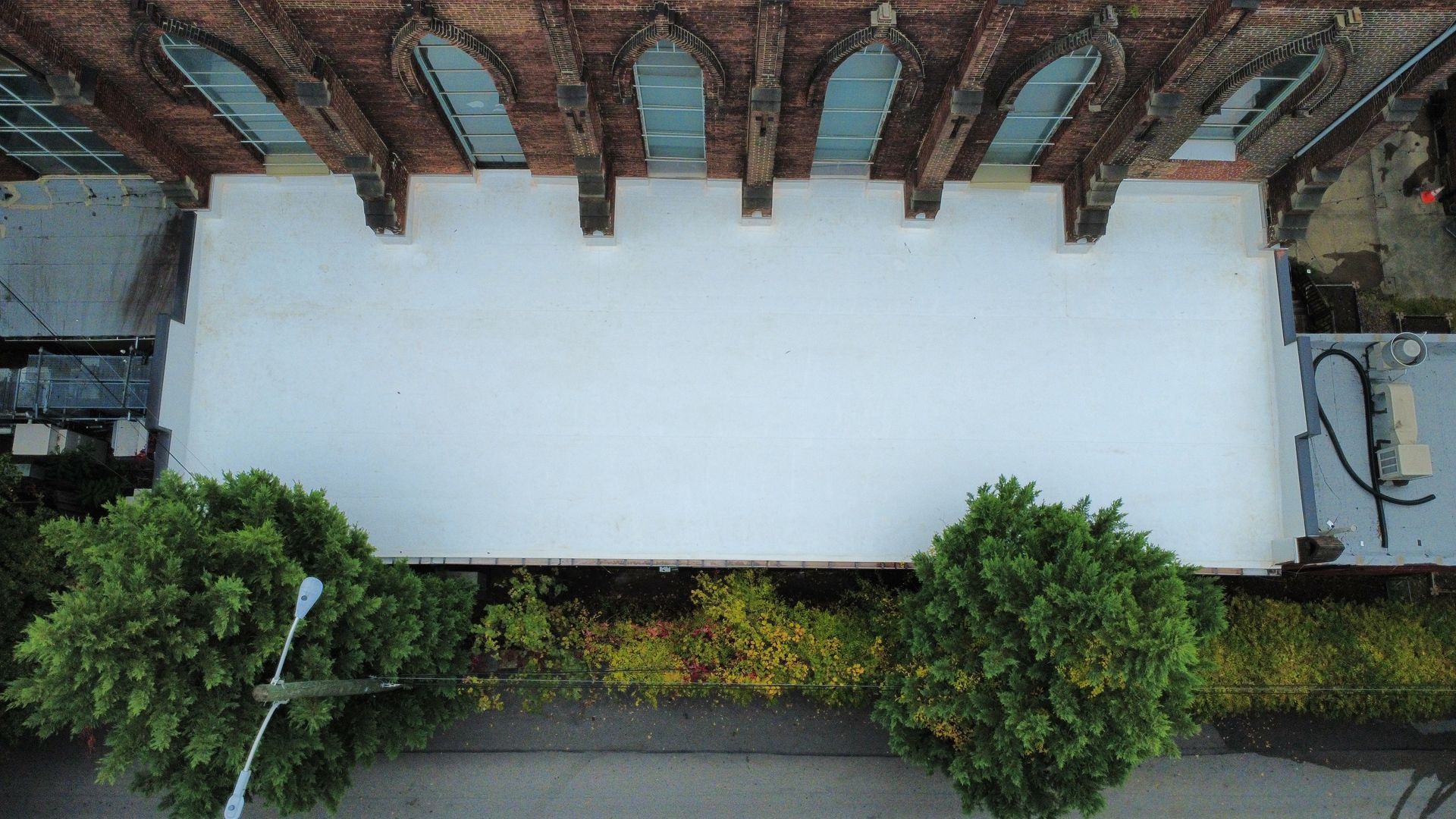 Overhead view of a white wall below a brick building with arched windows. Green bushes and a street lamp in the foreground.