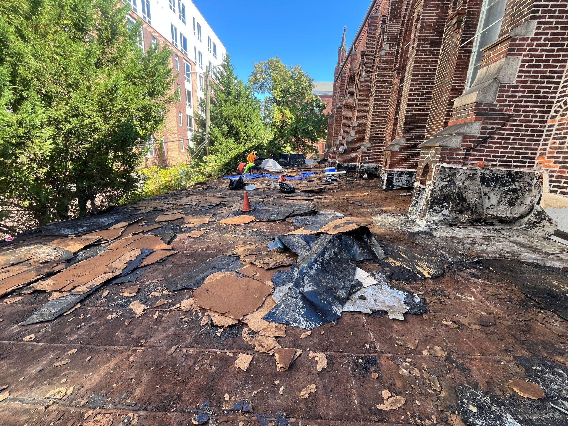 Damaged roof of a brick building. Black, peeling asphalt is exposed. Construction equipment and a person in background.