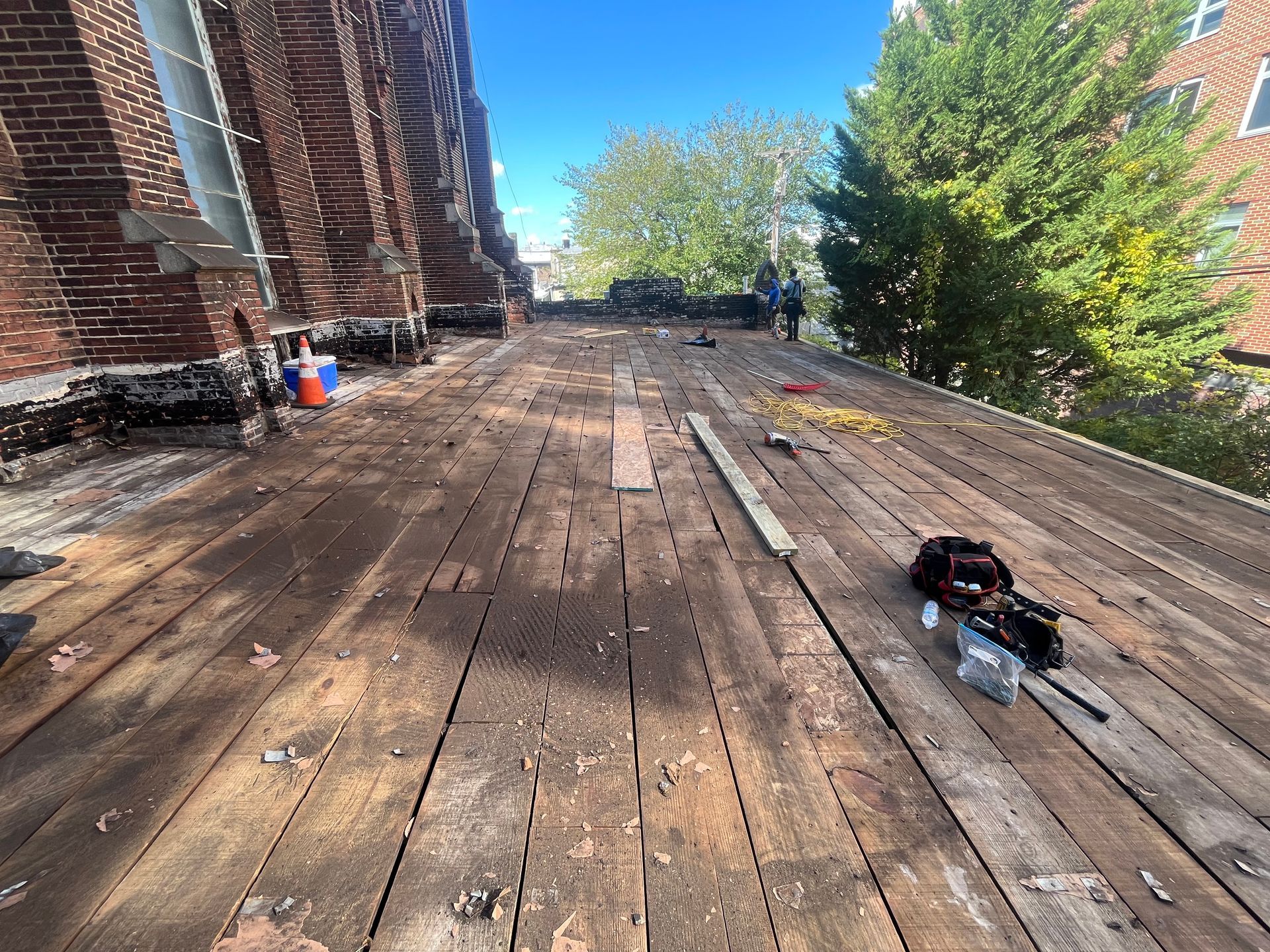 View of a roof with exposed wooden planks and construction materials, two people in the background, blue sky.