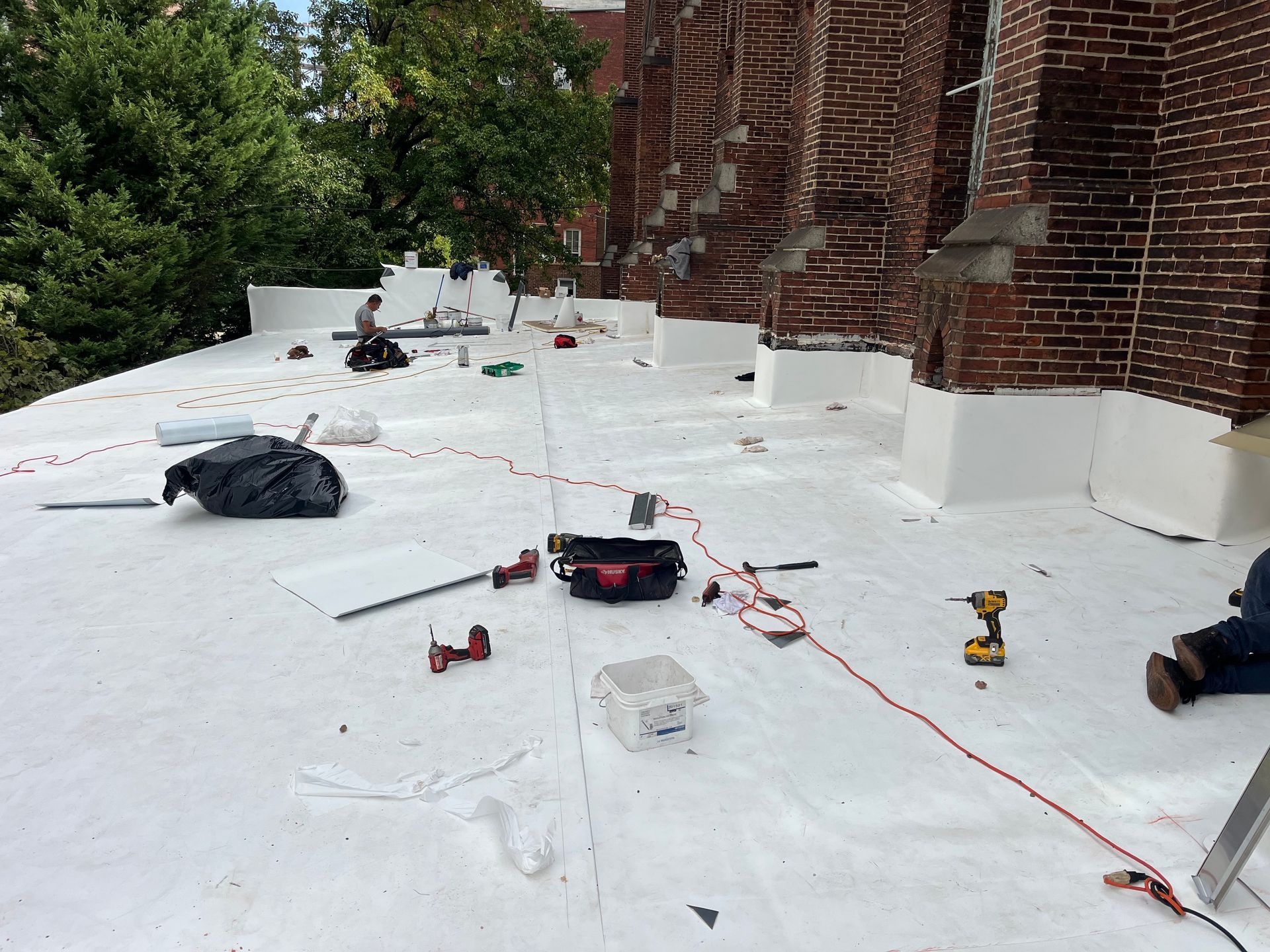 Workers on a white roof with tools. A brick building and trees are in the background.