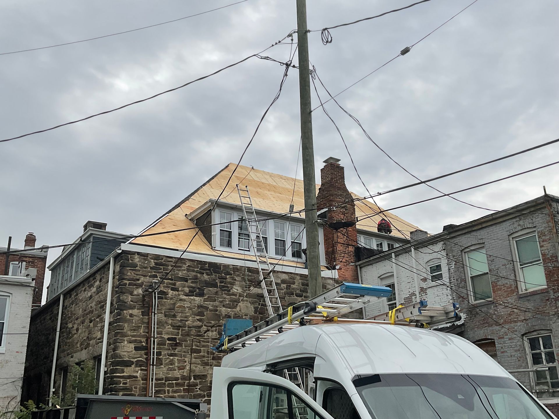 Roofing work on a house. Wooden roof, brick chimney, power lines, and a van with a ladder are visible.