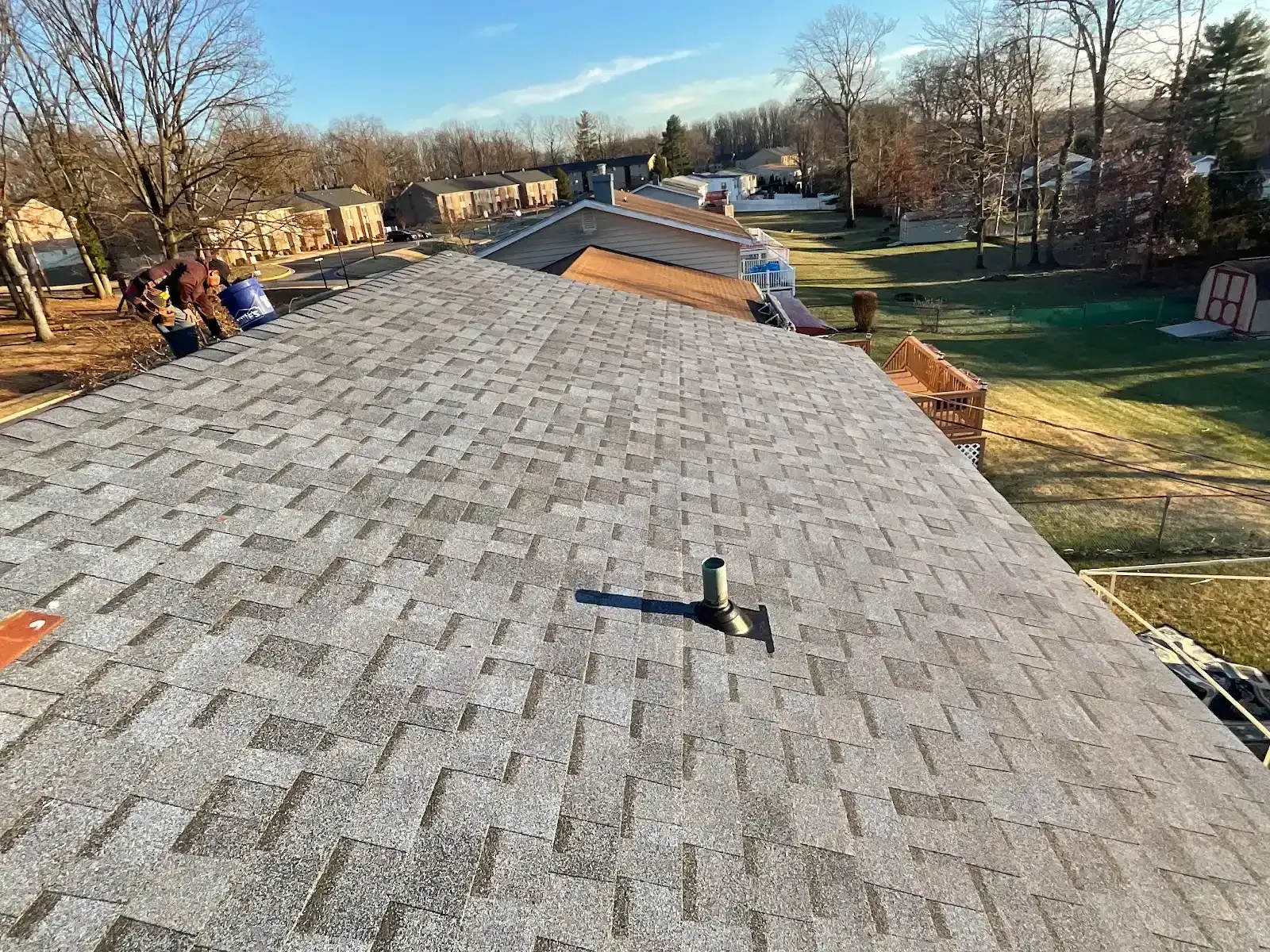 Gray shingle roof, view from above. A black vent pipe is on the roof. Construction workers on the left.
