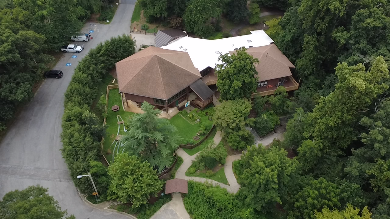 Overhead view of a building with a large hexagonal roof, surrounded by trees and a winding pathway.
