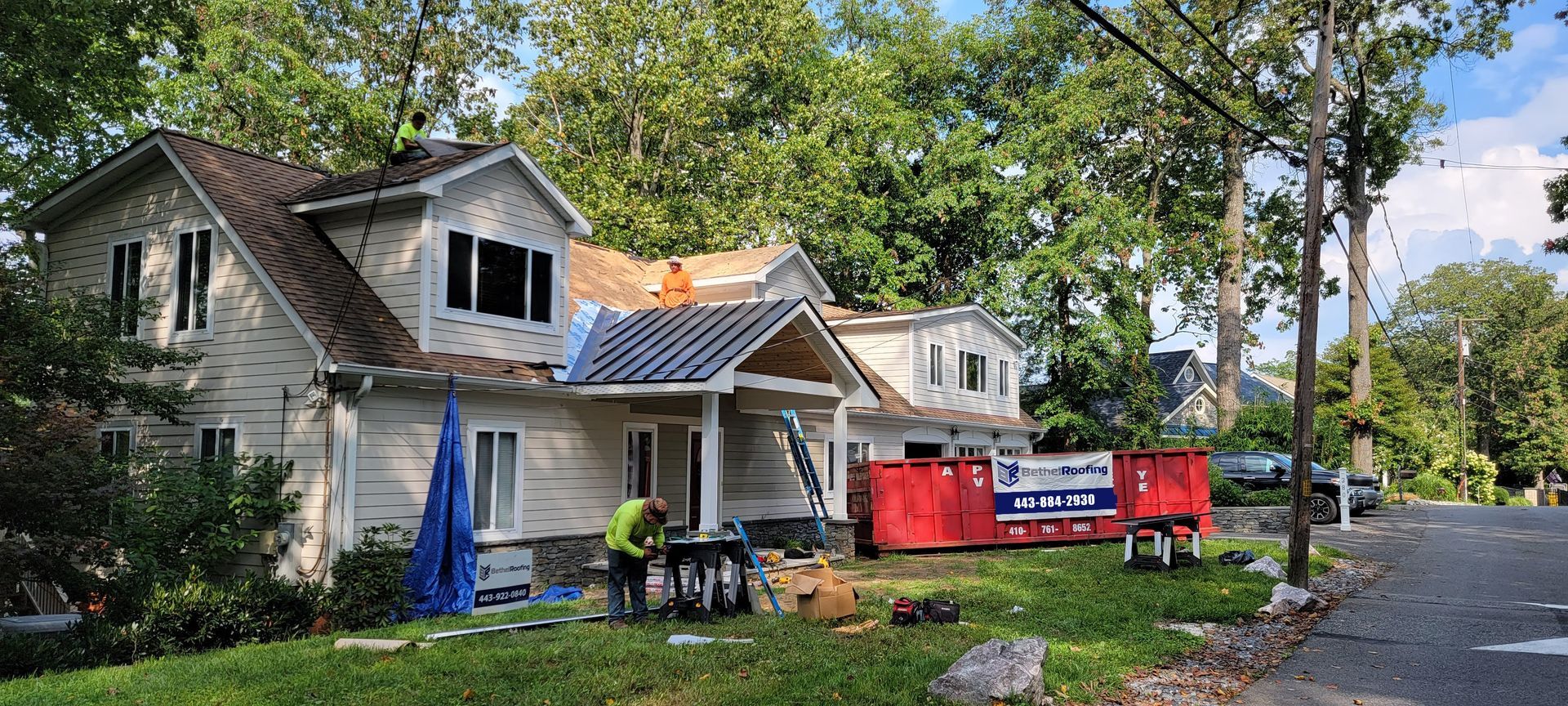 House under construction with workers, trees, and red dumpster in yard.