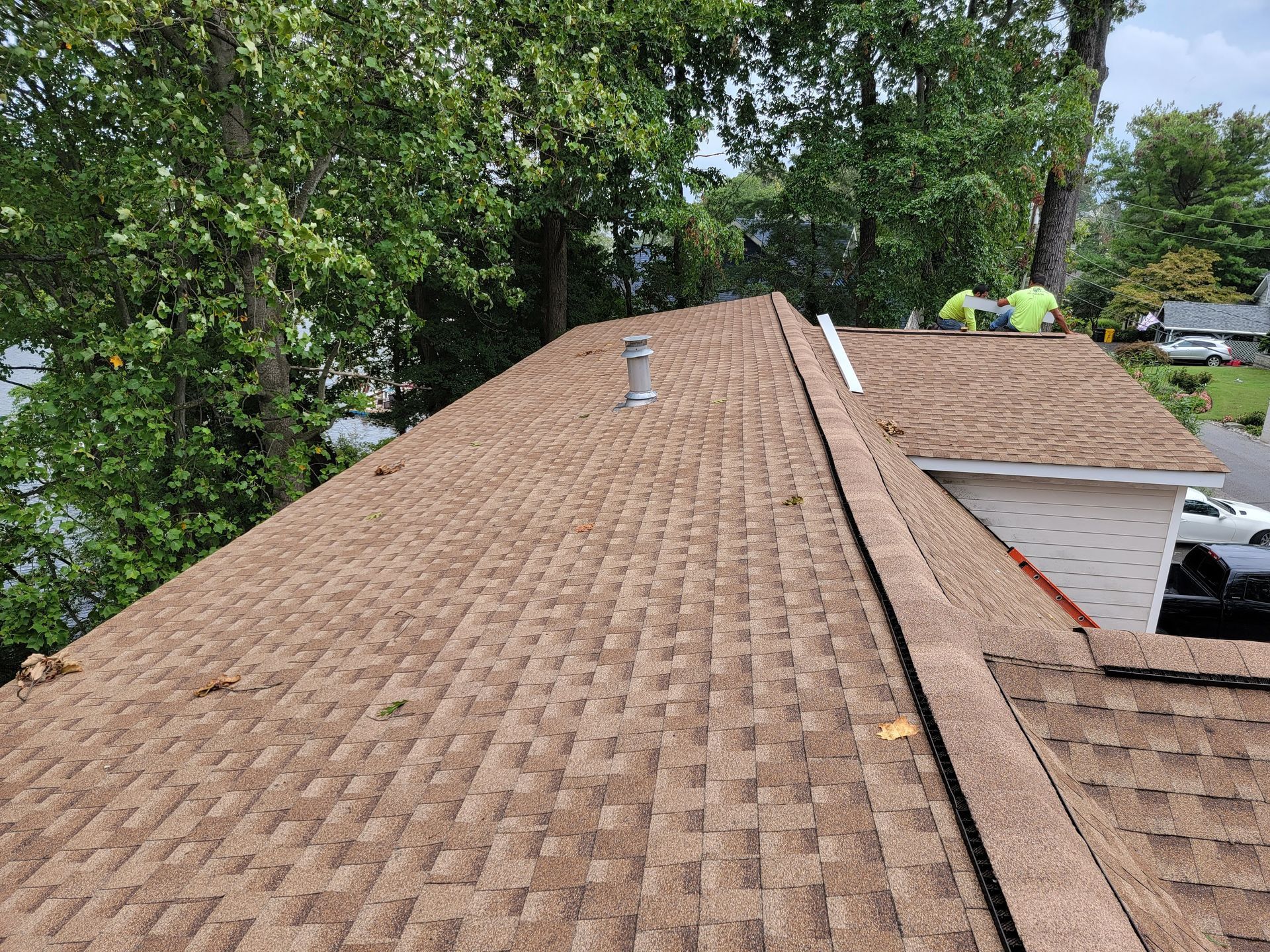 Brown shingle roof with two workers in safety vests. Trees in the background. Sunny day.