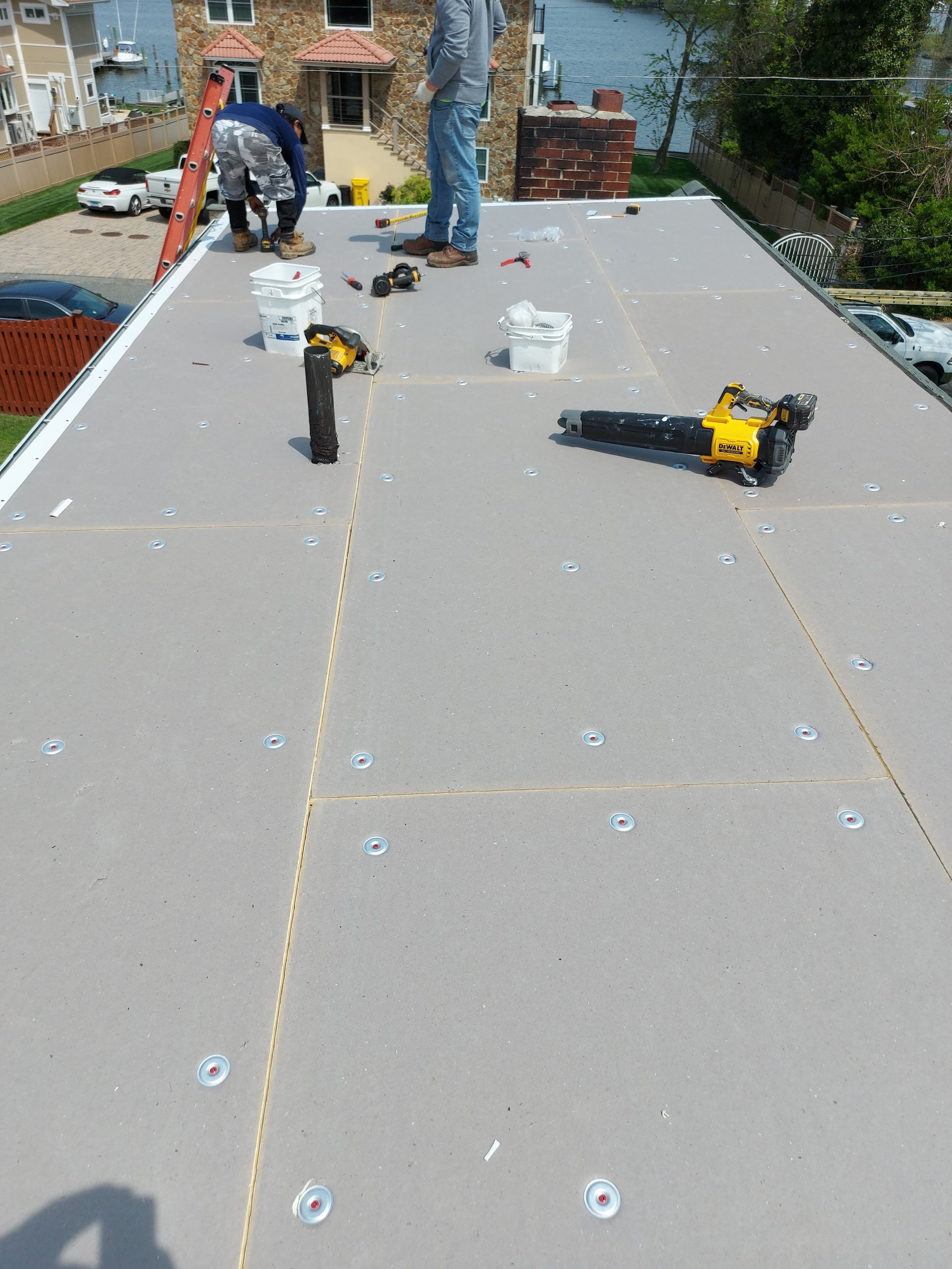 Workers installing roofing on a flat roof. Gray material, tools, buckets, and a house are visible.