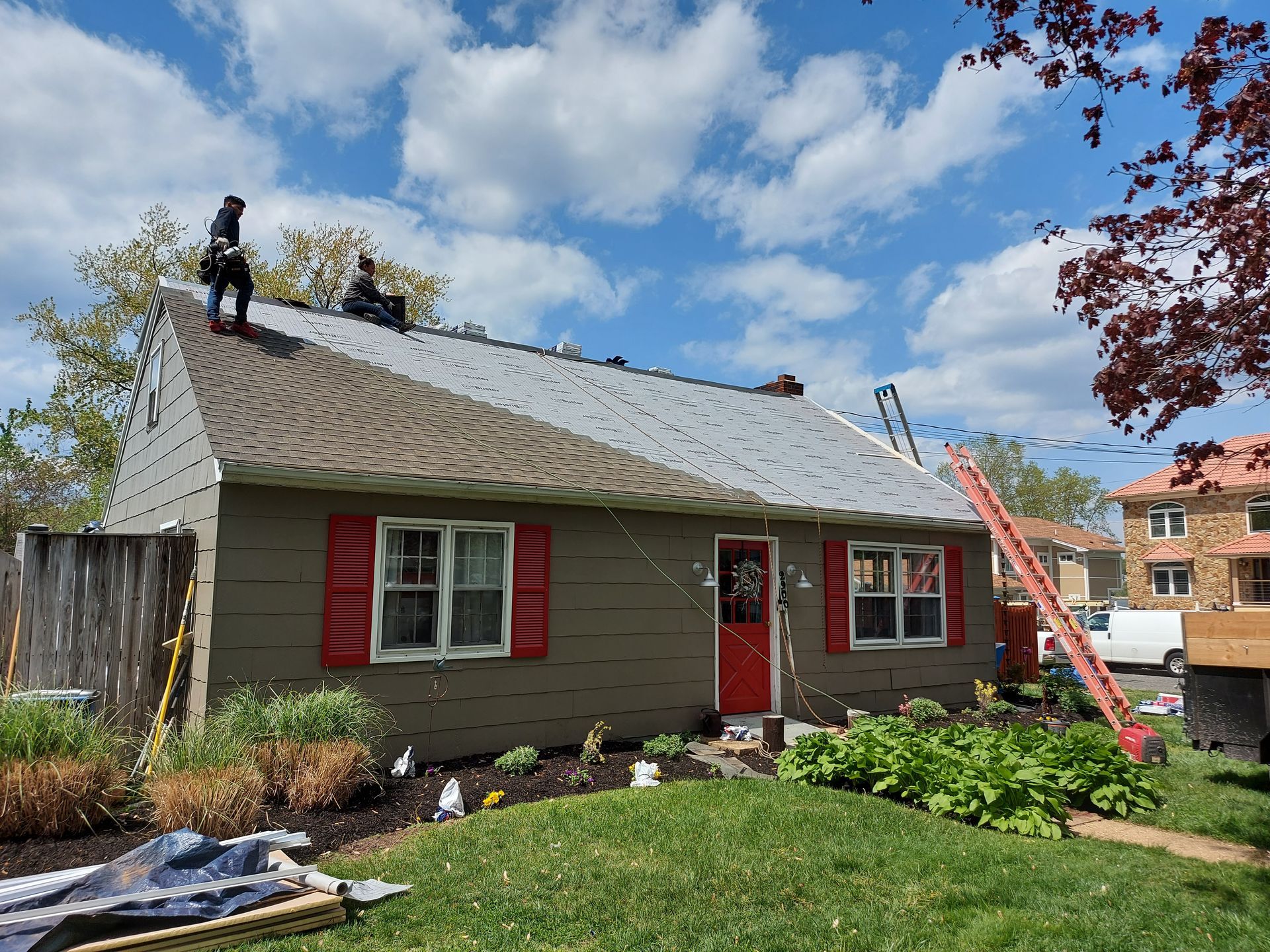 Roofers on a gray and brown shingled roof, red door, red shutters, blue sky, ladder.