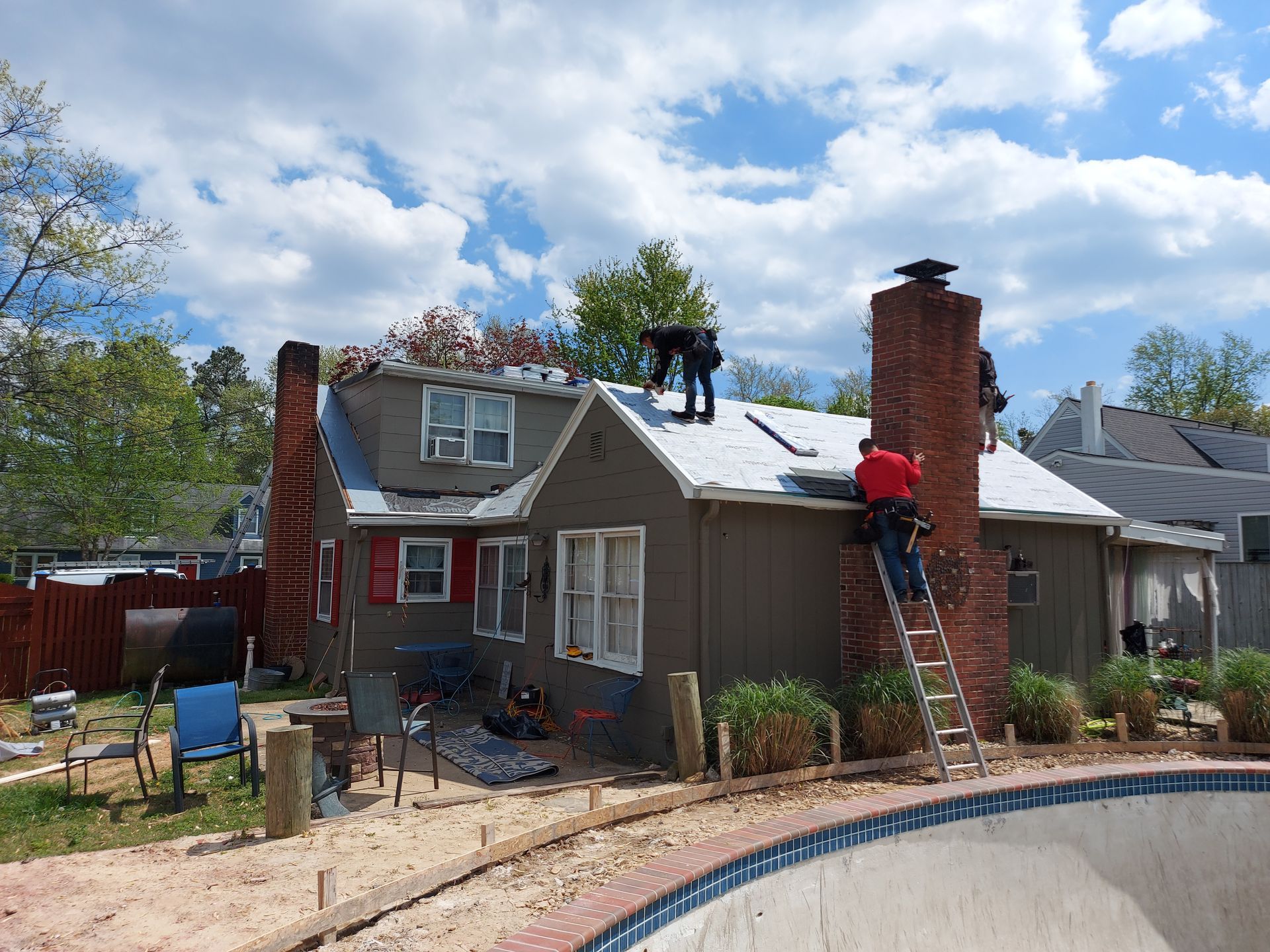 Workers on a roof replacing shingles of a house, with a ladder and chimney in view.