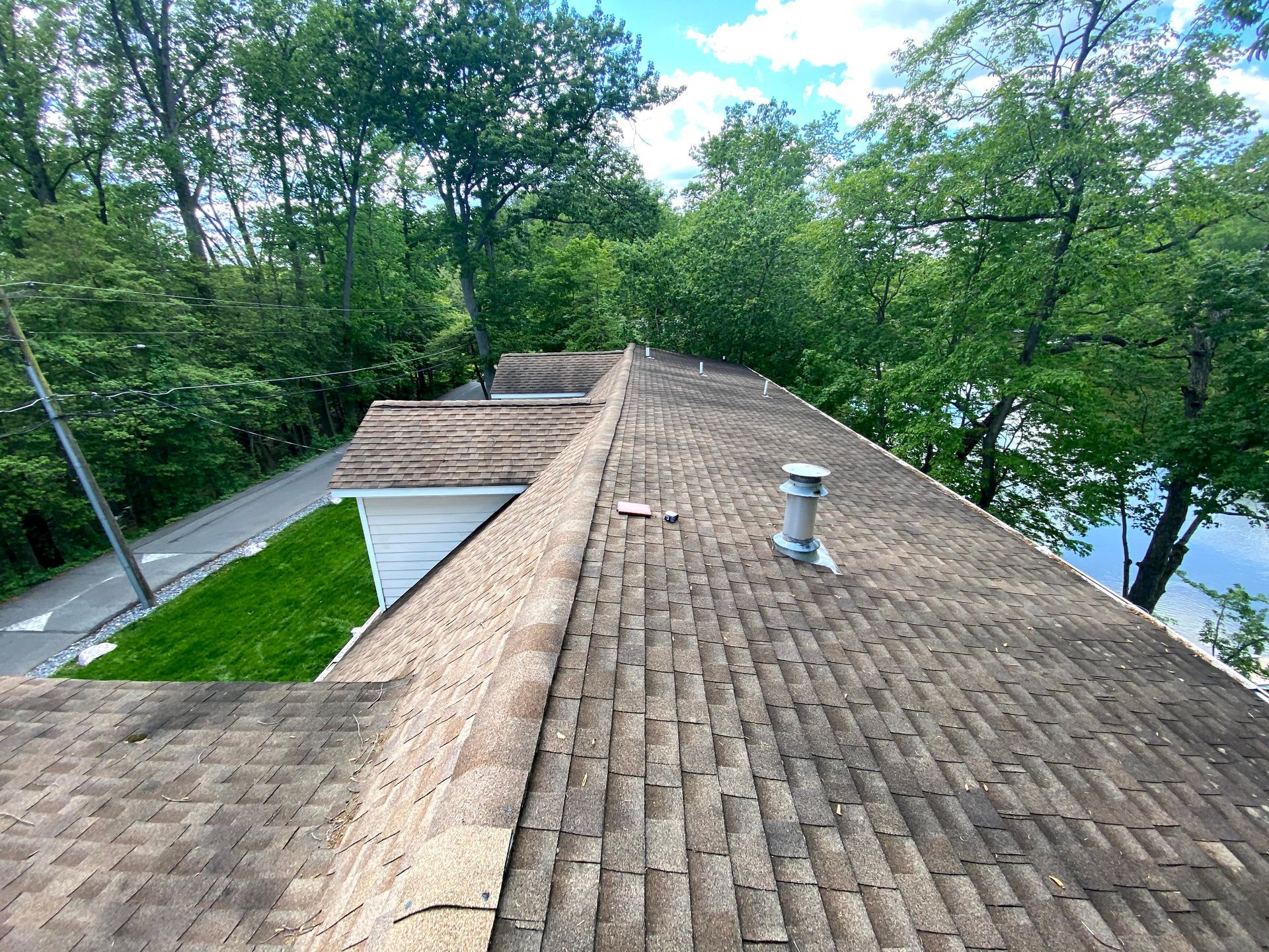 Brown shingle roof with a vent, surrounded by green trees and a visible road.