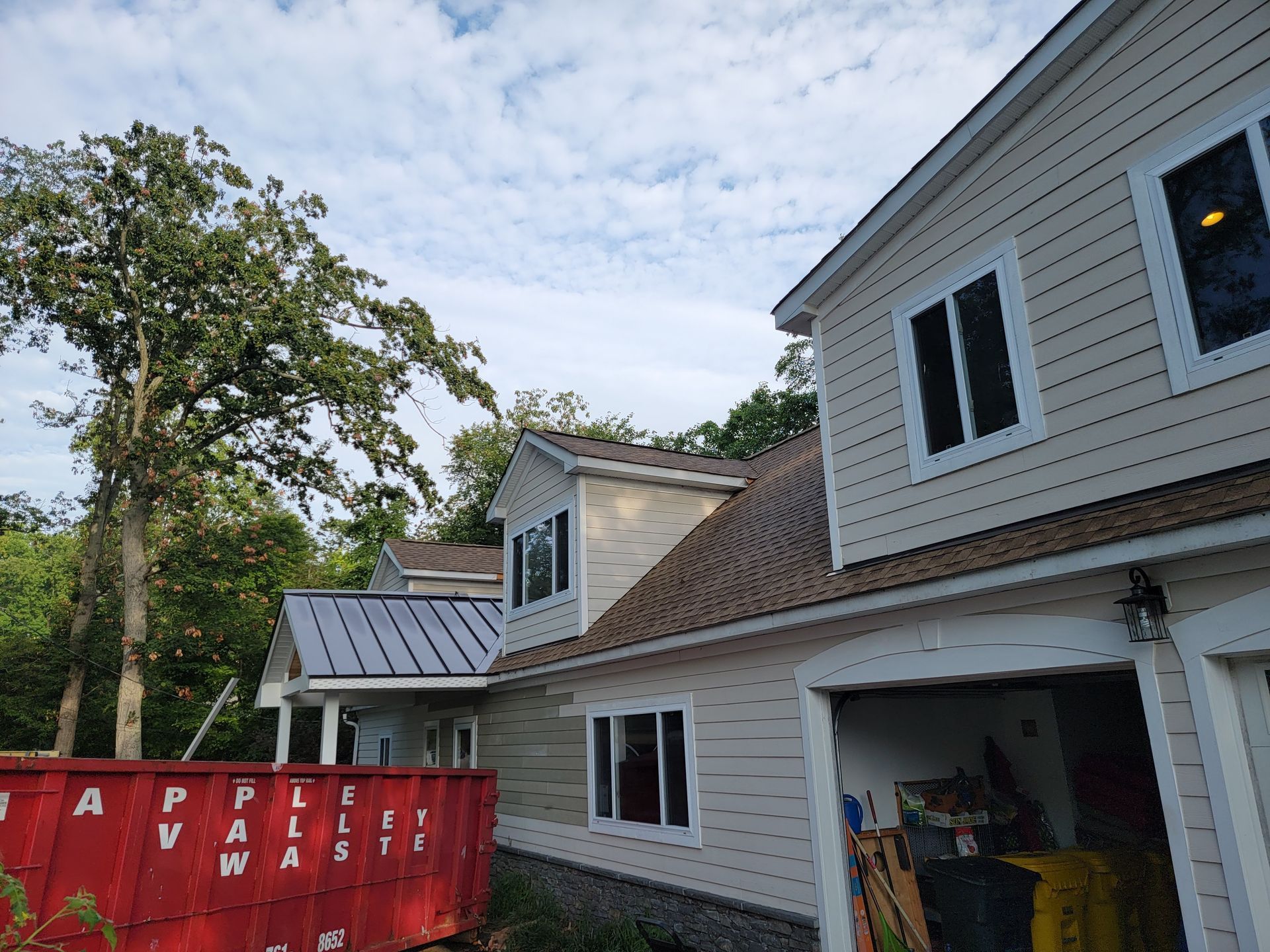Beige house with a brown roof and white trim, surrounded by trees. A red dumpster is visible.