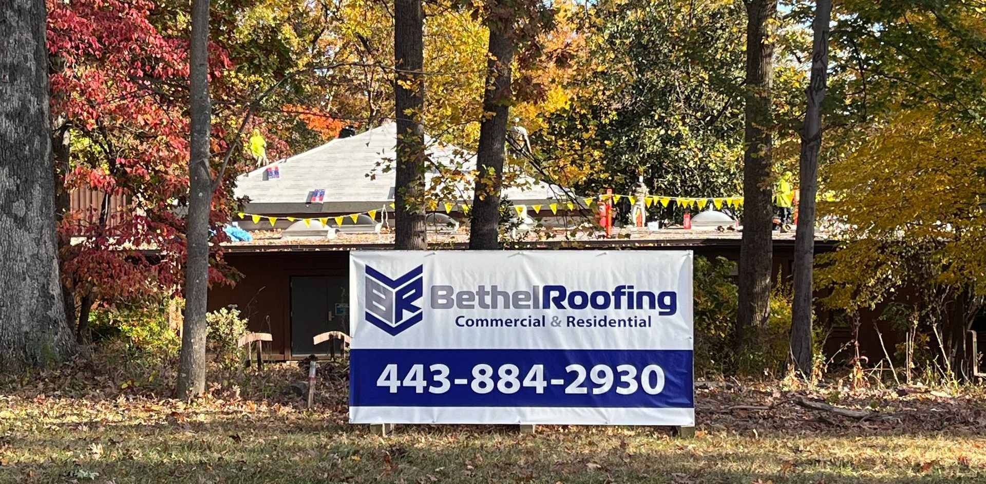 Sign for Bethel Roofing with a phone number, in front of a house, trees, and autumn foliage.