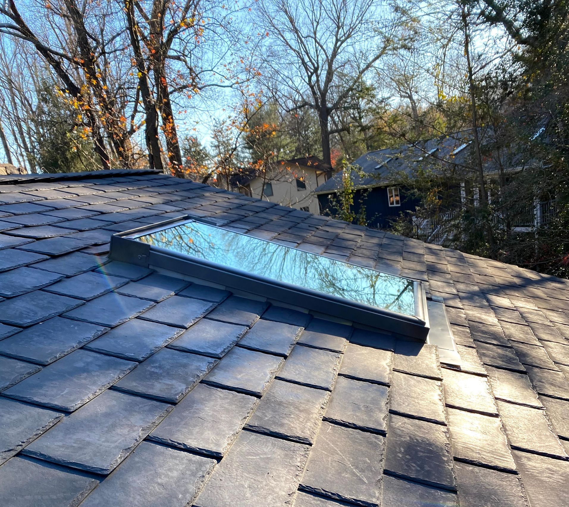 Skylight on a dark gray shingle roof. Trees and houses in the background on a sunny day.
