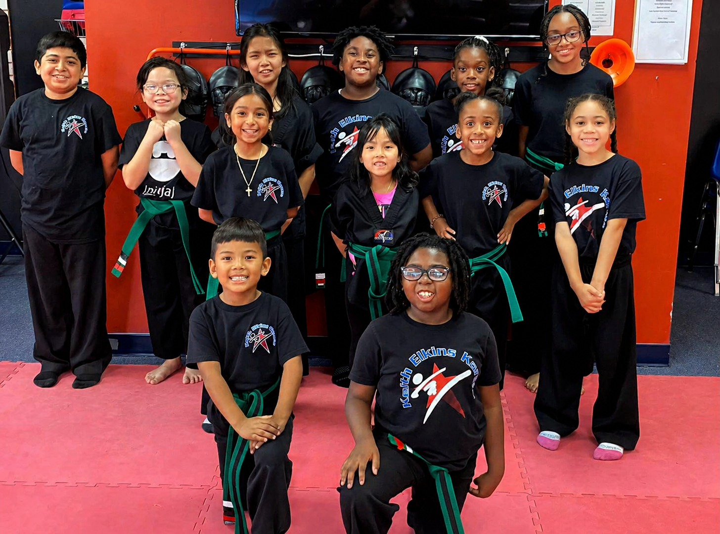 Five people in jiu-jitsu uniforms on a blue mat, including a man with a red belt, posing in a gym.