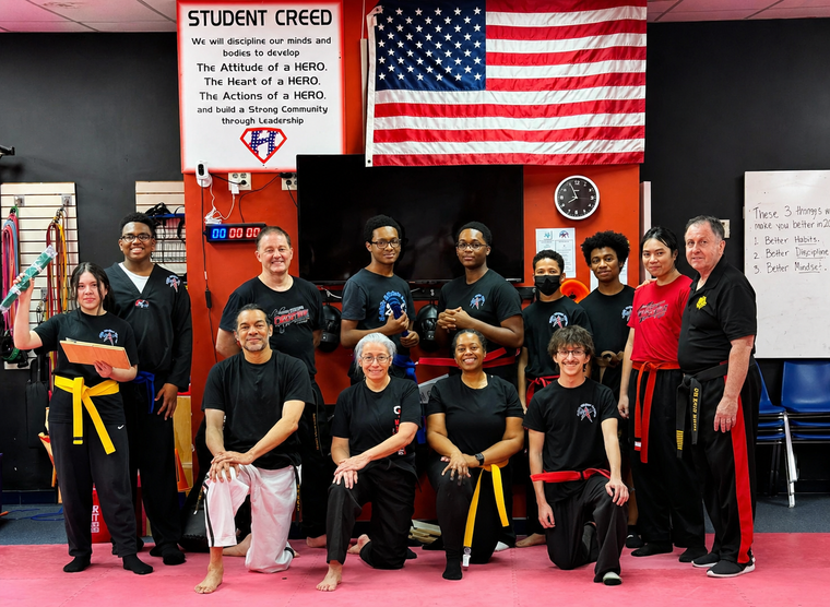 Family in Jiu-Jitsu gis poses. Indoors, blue mats, a smiling woman in the middle, around a seated man with a beard.