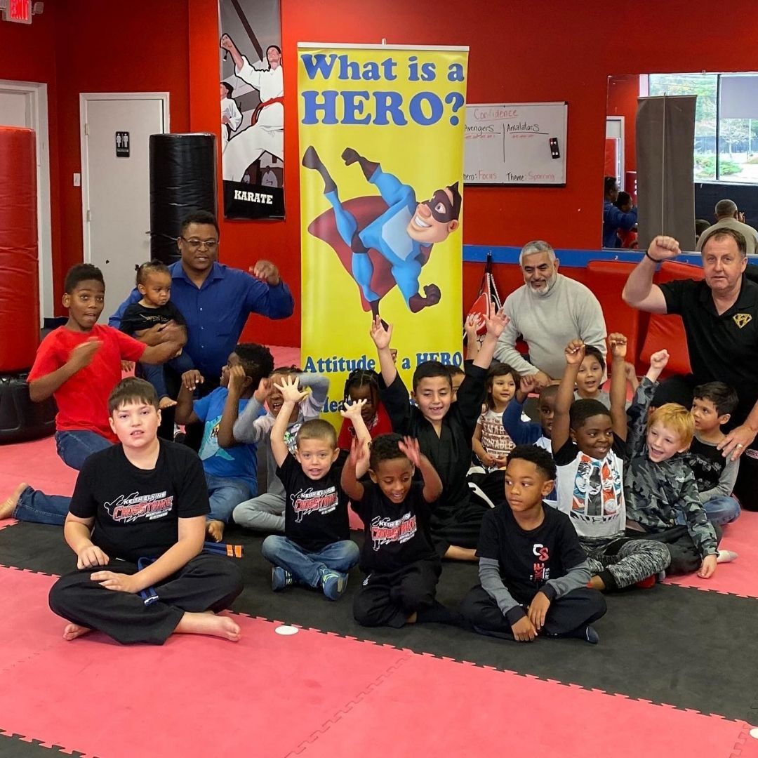 Two children in black and white martial arts uniforms practice a chokehold on a mat. One child smiles.