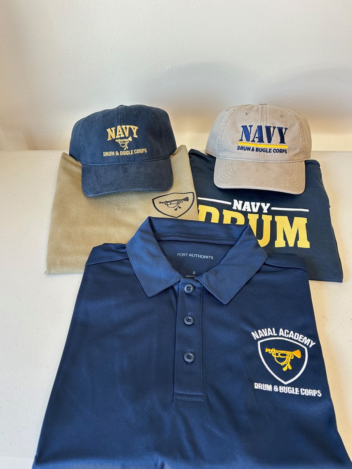A navy blue polo shirt, a tan shirt, and two Navy-branded baseball caps arranged on a light surface.
