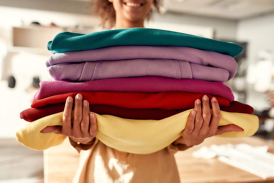 A smiling person holding a stack of colorful folded sweaters in a laundry setting.