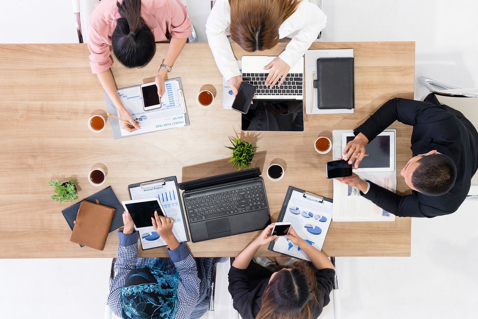 Overhead view of five people sitting around a wooden conference table, using laptops, tablets, and phones to review papers.