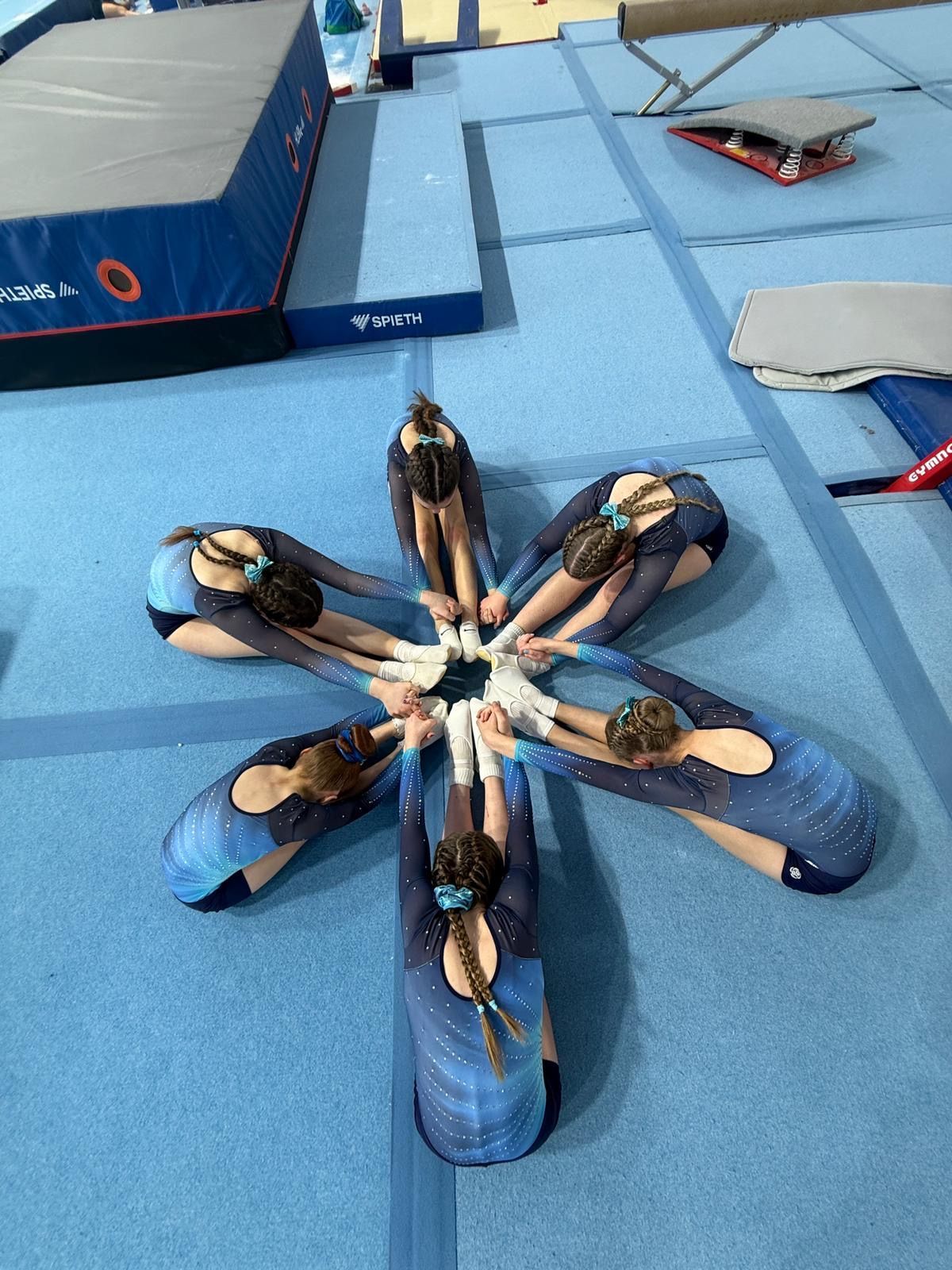 Children lying in a star shape on blue gym mats, with arms extended toward the center.