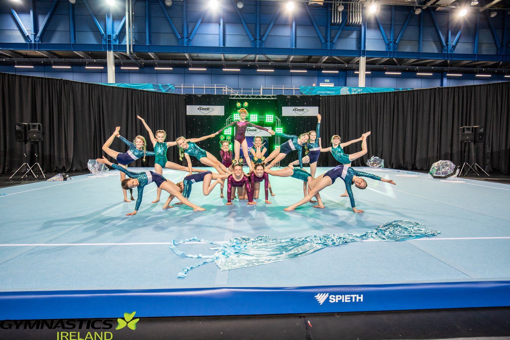 Gymnastics team posing on a blue floor mat in an indoor arena, with arms raised and a green-lit stage backdrop.